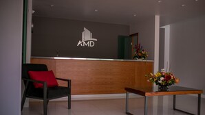 Un área de recepción con un mostrador de madera, una silla negra con cojín rojo y un jarrón con flores.