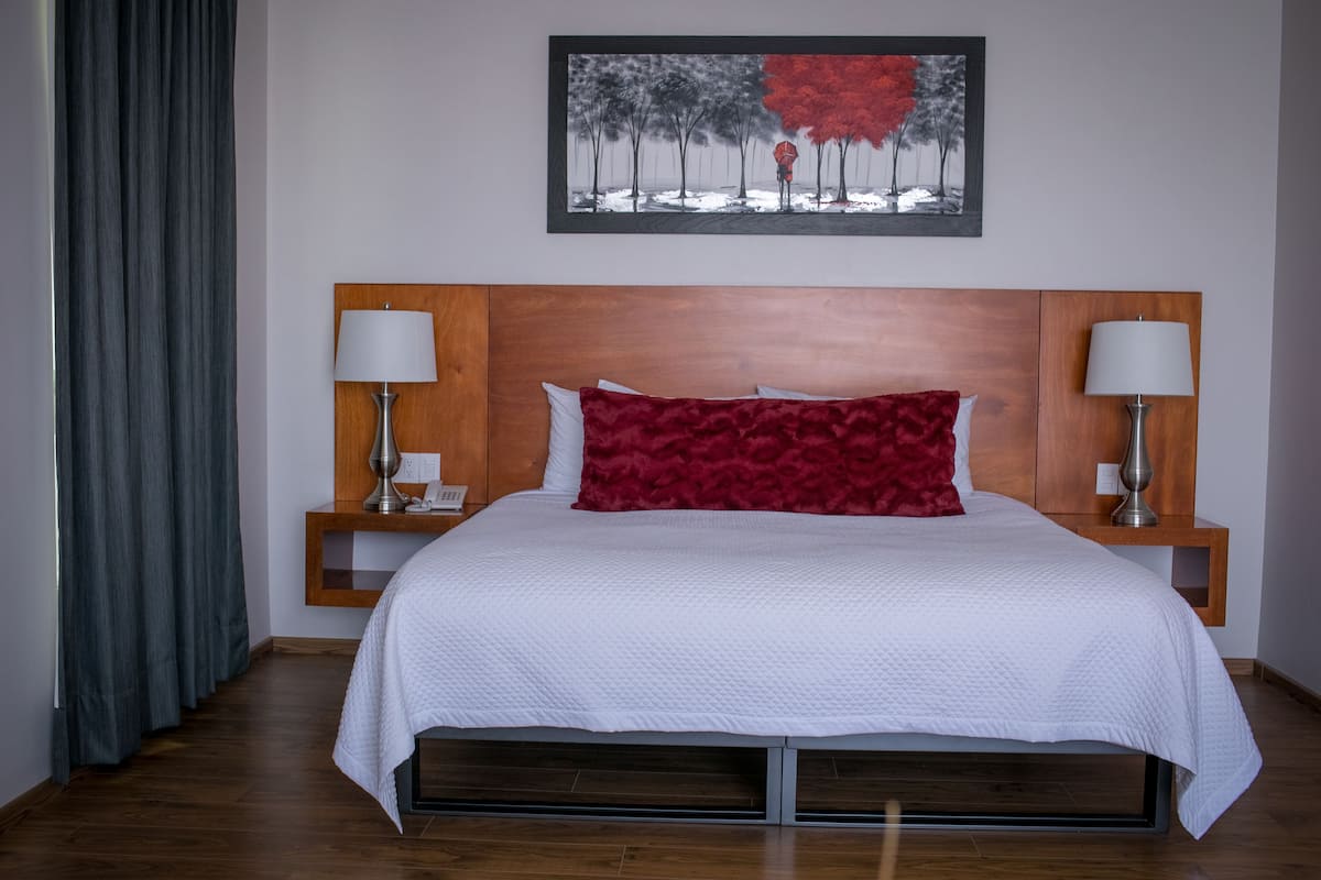 Un lit bien fait, recouvert d’une couette blanche et agrémenté d’un oreiller rouge, est encadré par deux tables de chevet sur lesquelles reposent des lampes. La pièce est dotée d’un plancher en bois et un tableau est accroché au-dessus du lit.