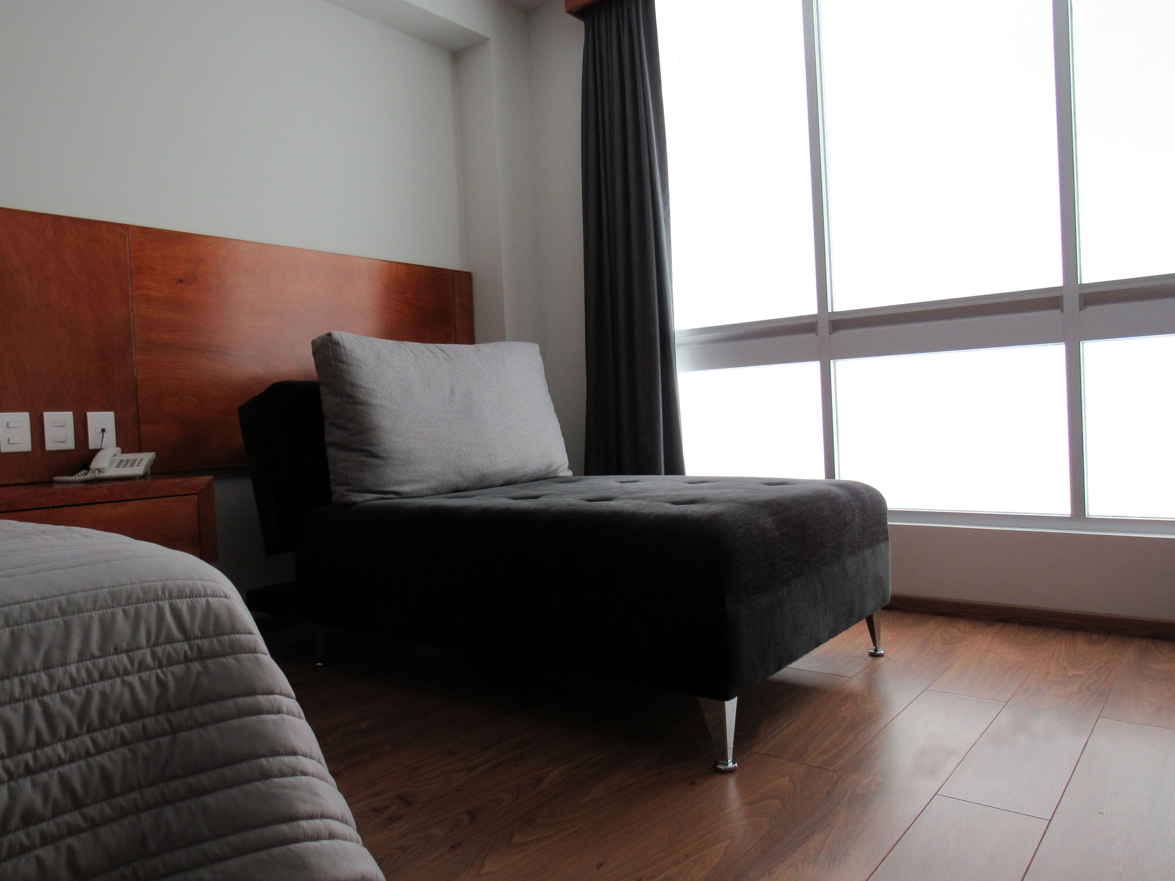 A hotel room with a wooden headboard, a single bed with a grey quilt, a small bedside table with a telephone, and a large window with curtains.