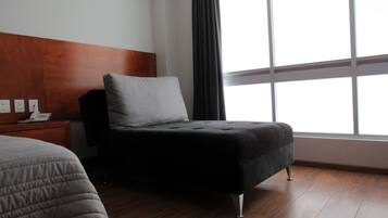 A hotel room with a wooden headboard, a single bed with a grey quilt, a small bedside table with a telephone, and a large window with curtains.