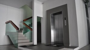 Un ascensor moderno con detalles metálicos, junto a una escalera con barandillas de vidrio y escalones de madera.