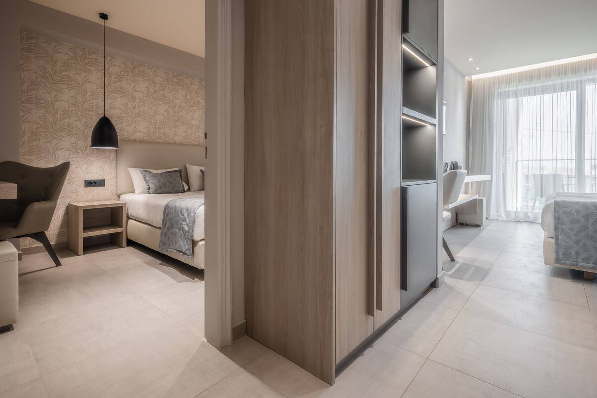Quarto de hotel moderno com cama, mesas de cabeceira, guarda-roupa e área de estar com uma cadeira e uma pequena mesa.