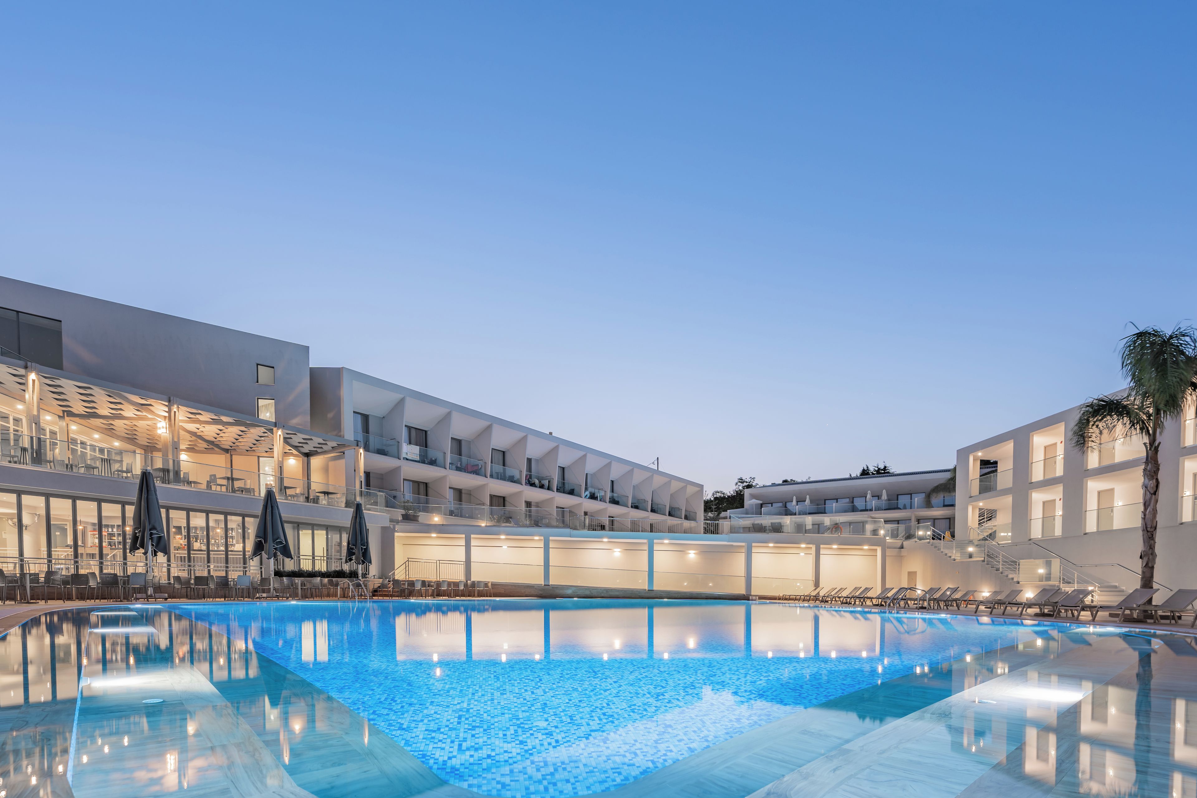 Un hotel moderno con piscina, mobiliario exterior y fachada luminosa.