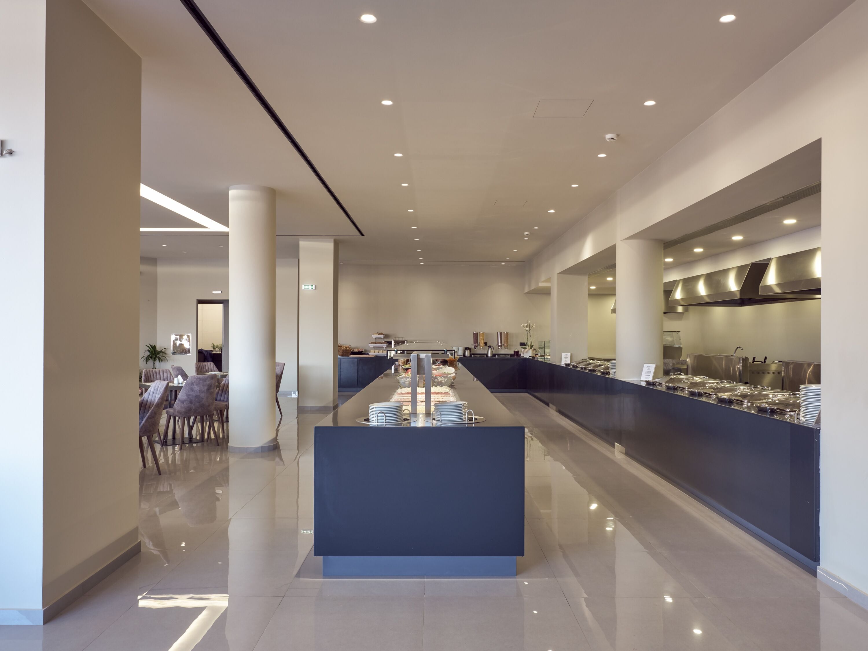 Um lobby de hotel moderno com uma recepção central, área de estar e uma cozinha visível ao fundo.