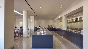 Um lobby de hotel moderno com uma recepção central, área de estar e uma cozinha visível ao fundo.