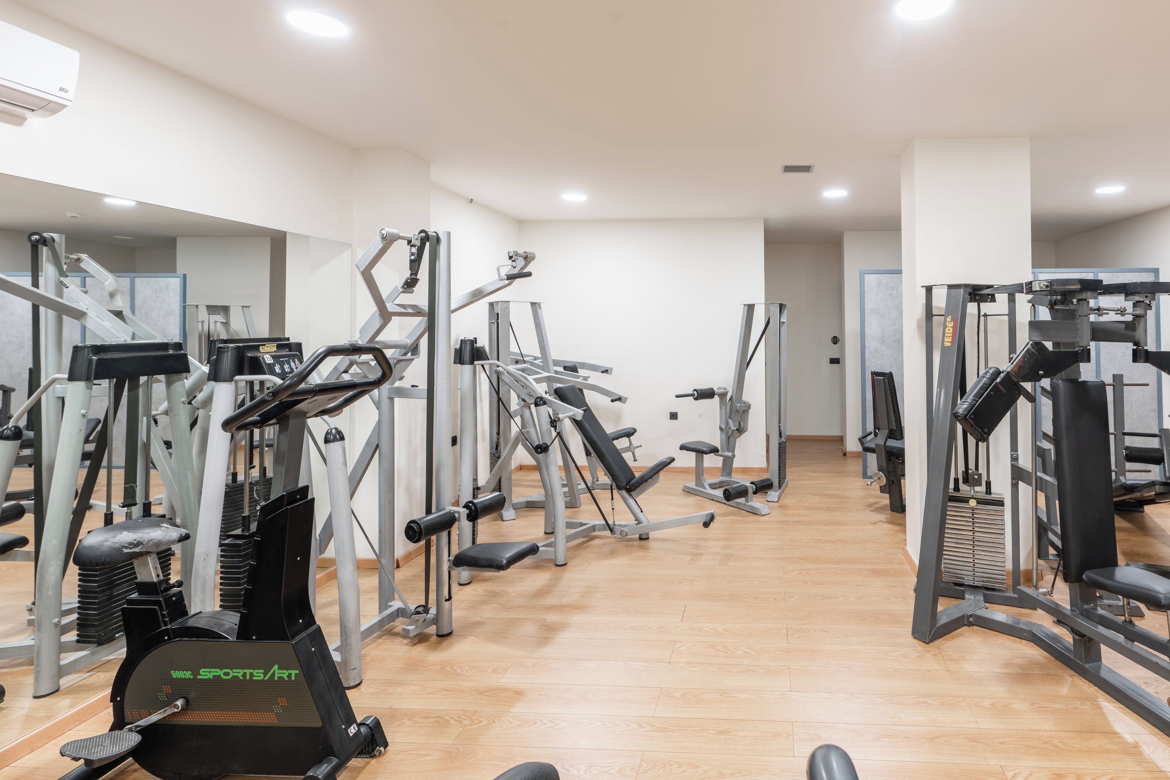 Een sportschool met diverse fitnessapparaten, een spiegelwand en houten vloeren.