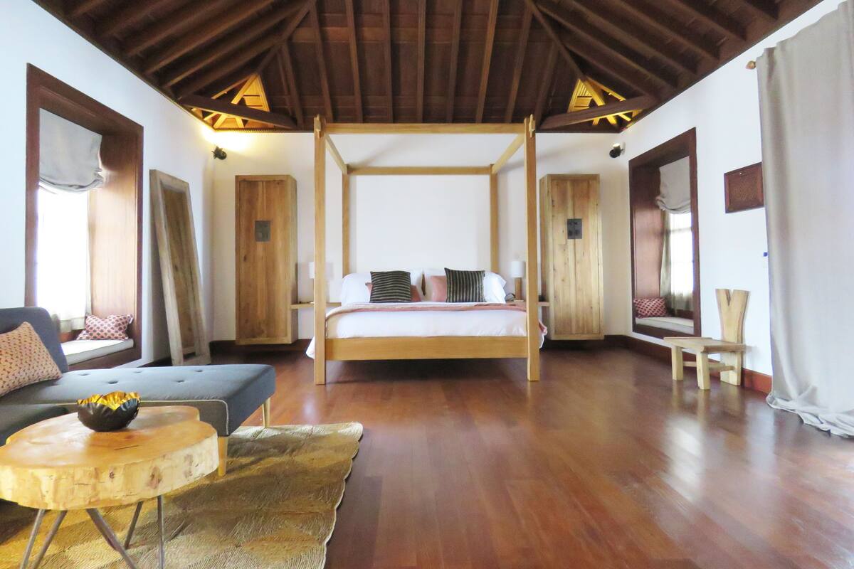 A spacious bedroom with a four-poster bed, wooden flooring, and a high ceiling.