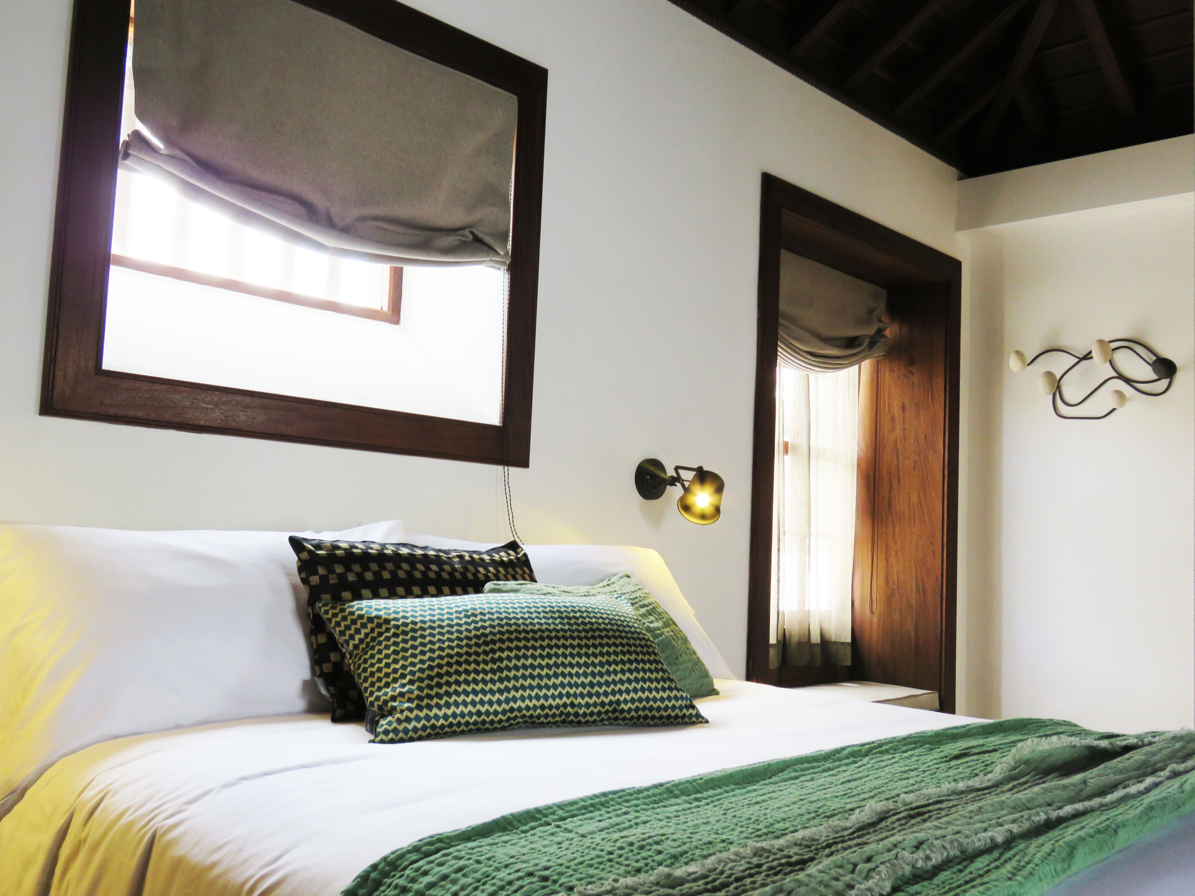 A bedroom with a bed, pillows, a bedside lamp, a window with a curtain, and a wall-mounted light fixture.