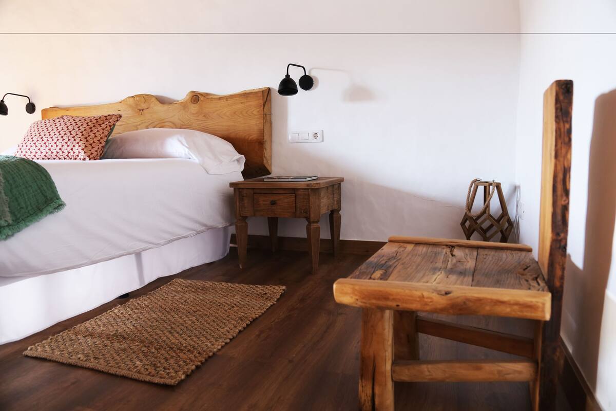 A bedroom with a wooden headboard, a bedside table with a lamp, a wooden chair, and a woven rug.