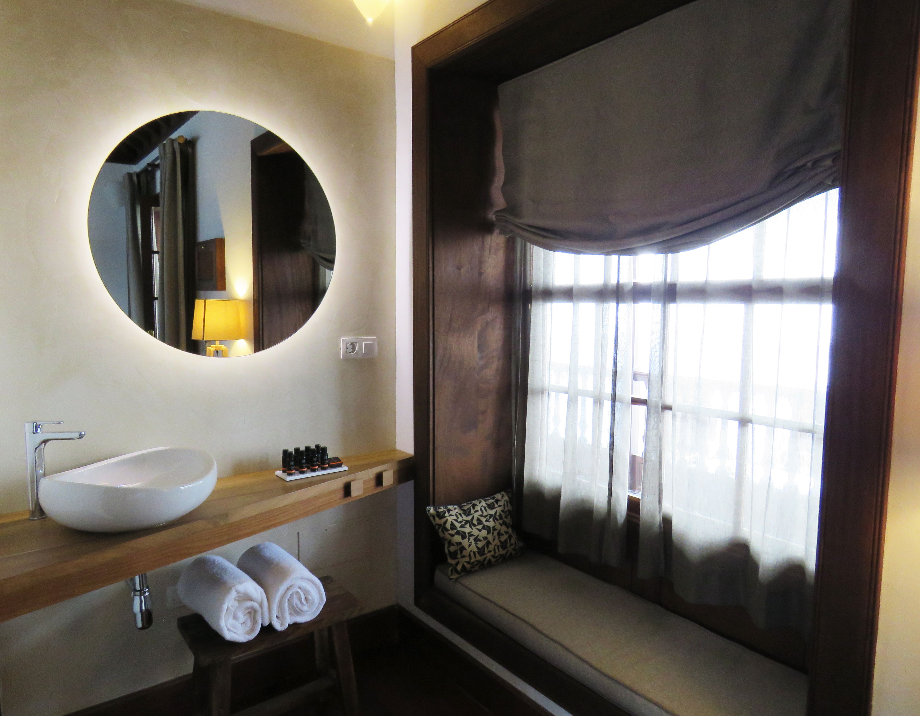 Un baño con un espejo redondo, un lavabo blanco, un mueble de tocador de madera y una ventana con cortinas.