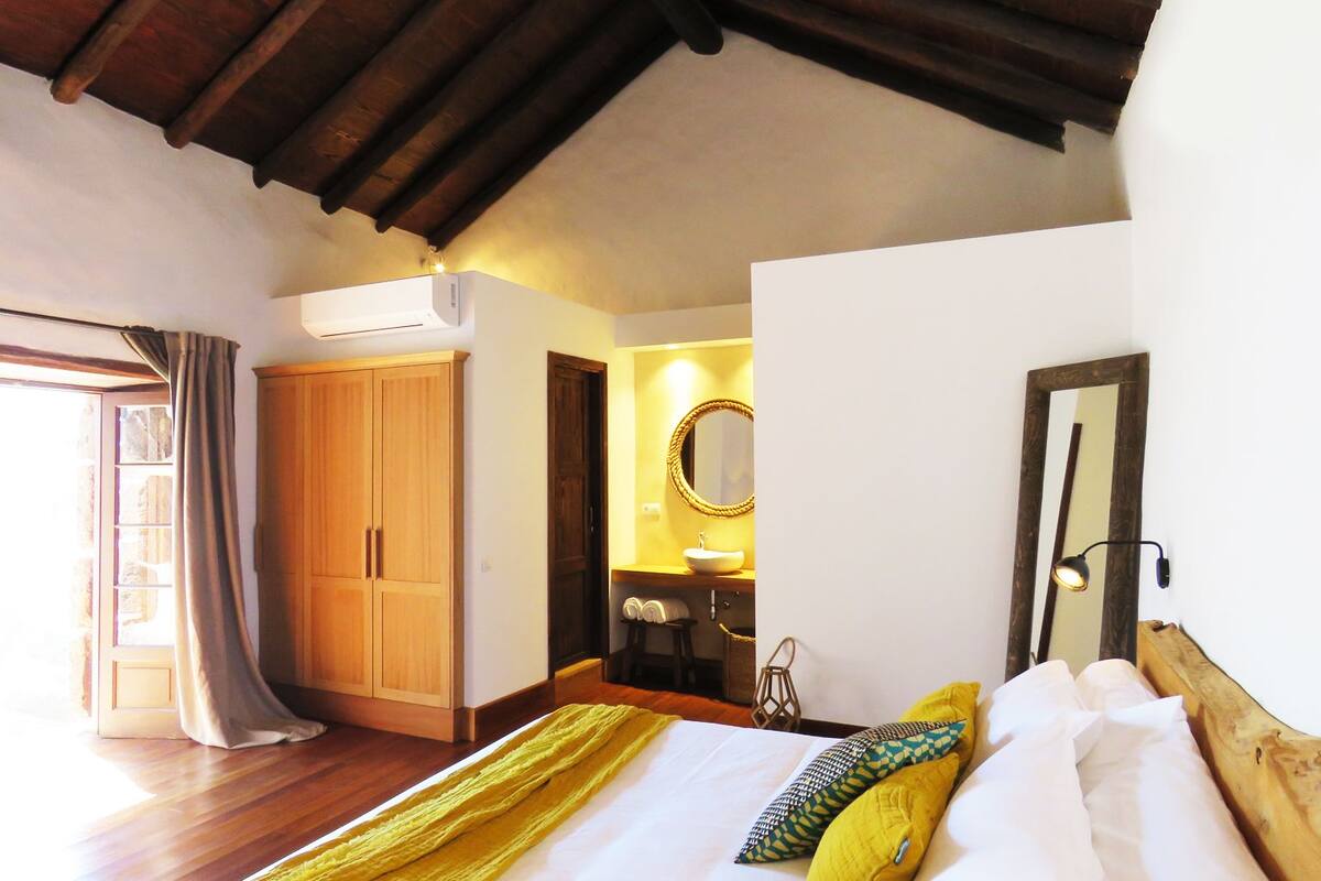 A bedroom with a wooden wardrobe, a bed with white and yellow bedding, a bedside table with a lamp, and a mirror on the wall.