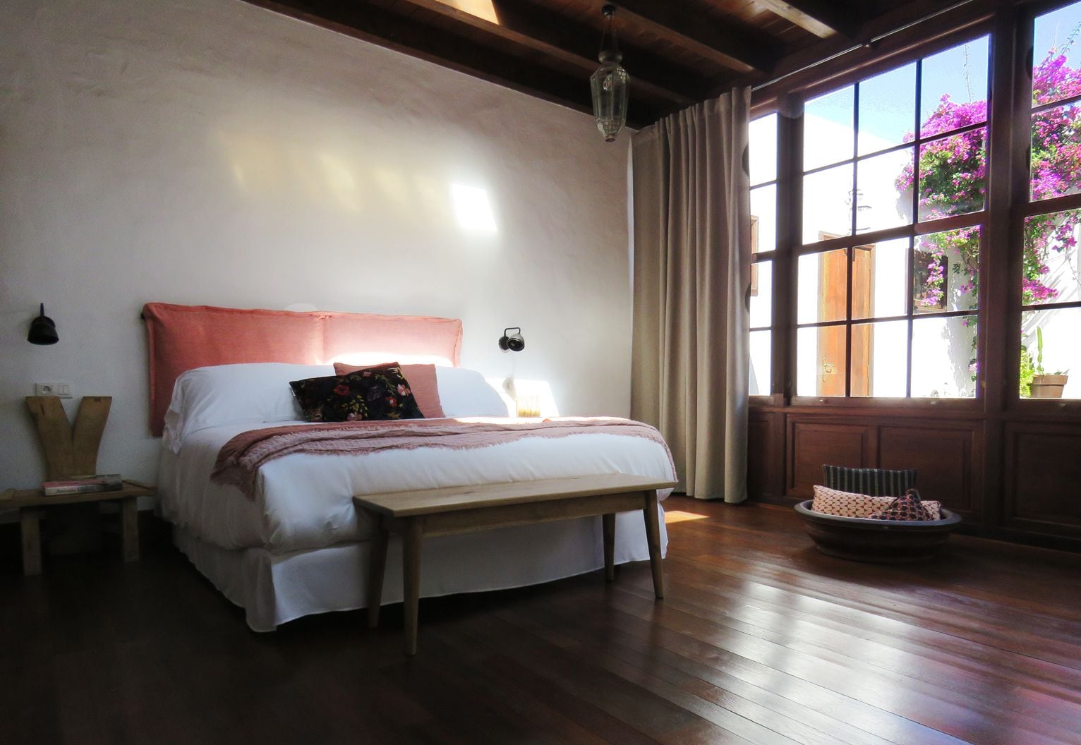 A bedroom with a large bed, wooden floor, and a window with a view of flowers.