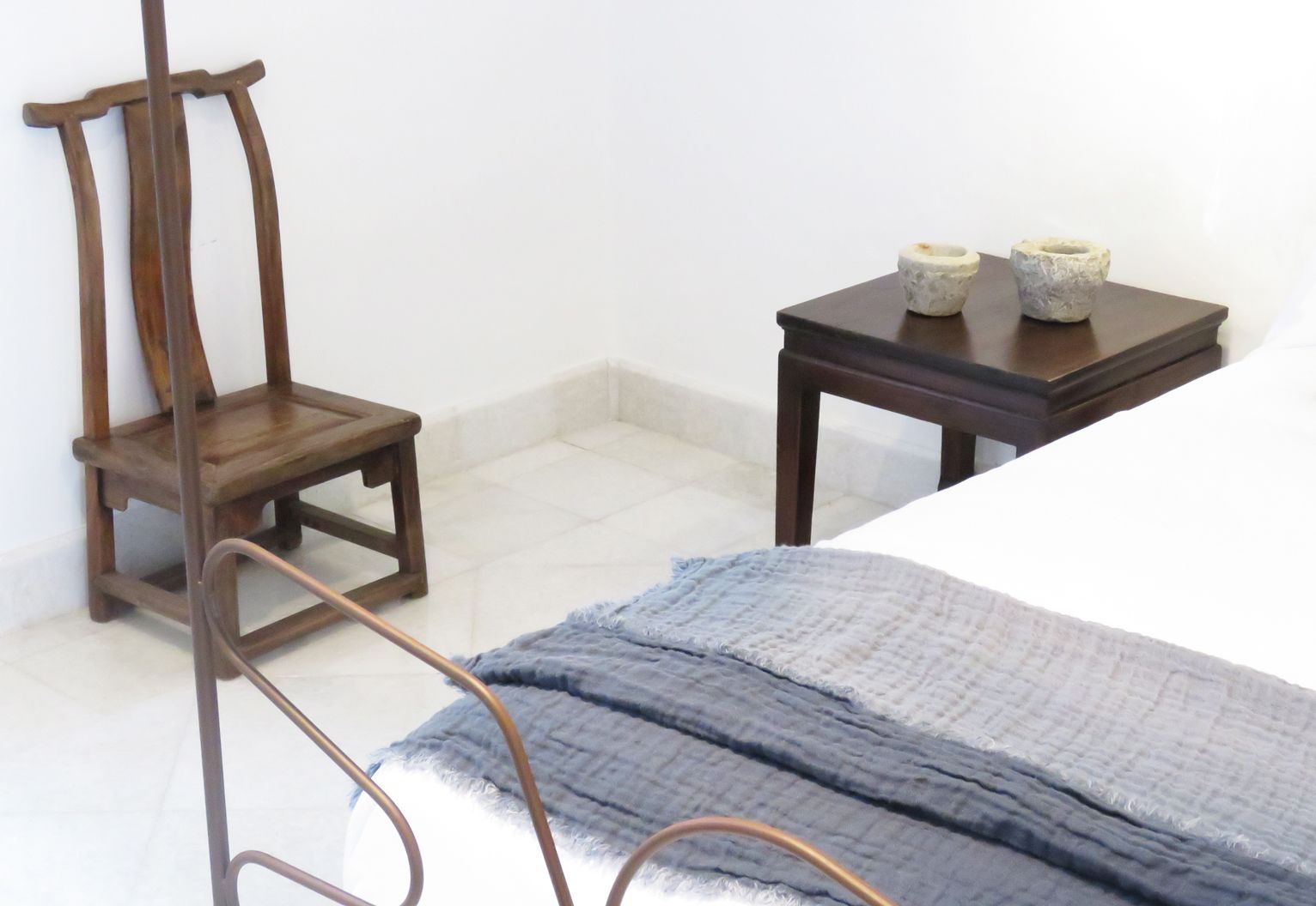 Un dormitorio con una cama, una silla de madera, una mesita con dos objetos decorativos y una manta azul.