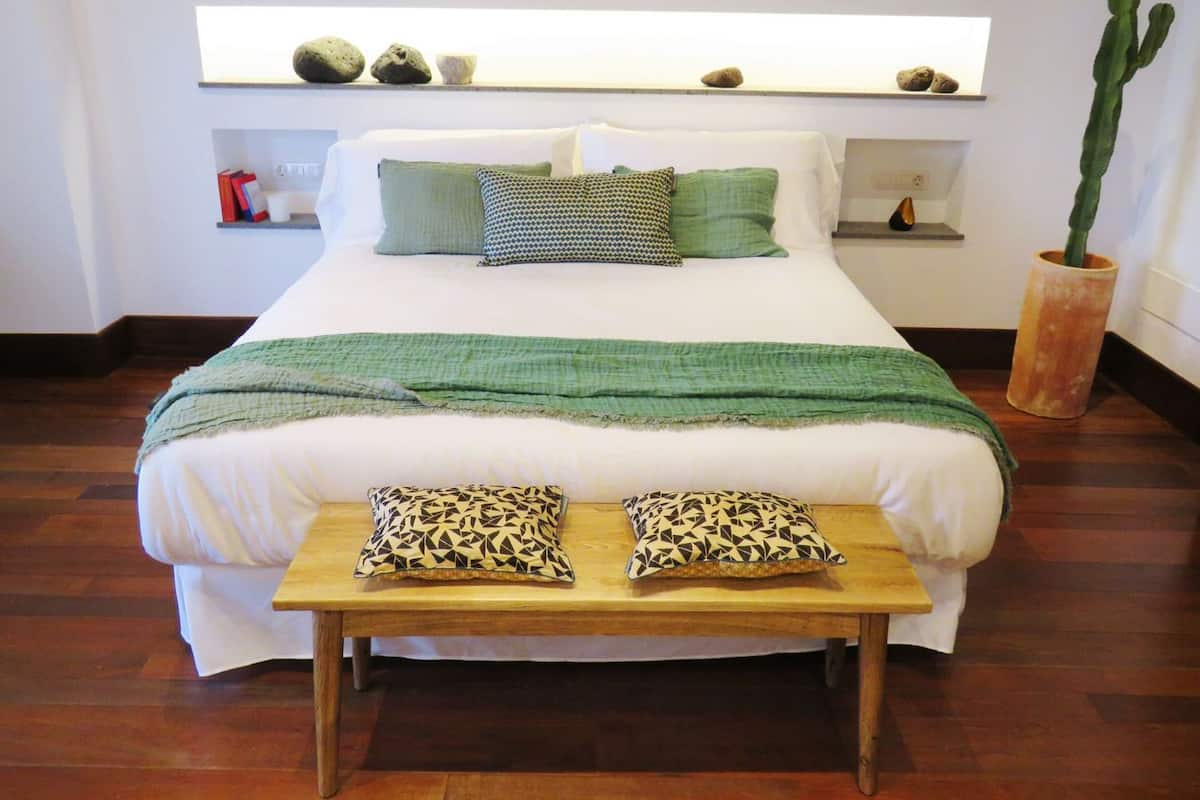 A neatly made bed with a green blanket and patterned pillows, a wooden bench with matching cushions, and a cactus in a pot on the floor.