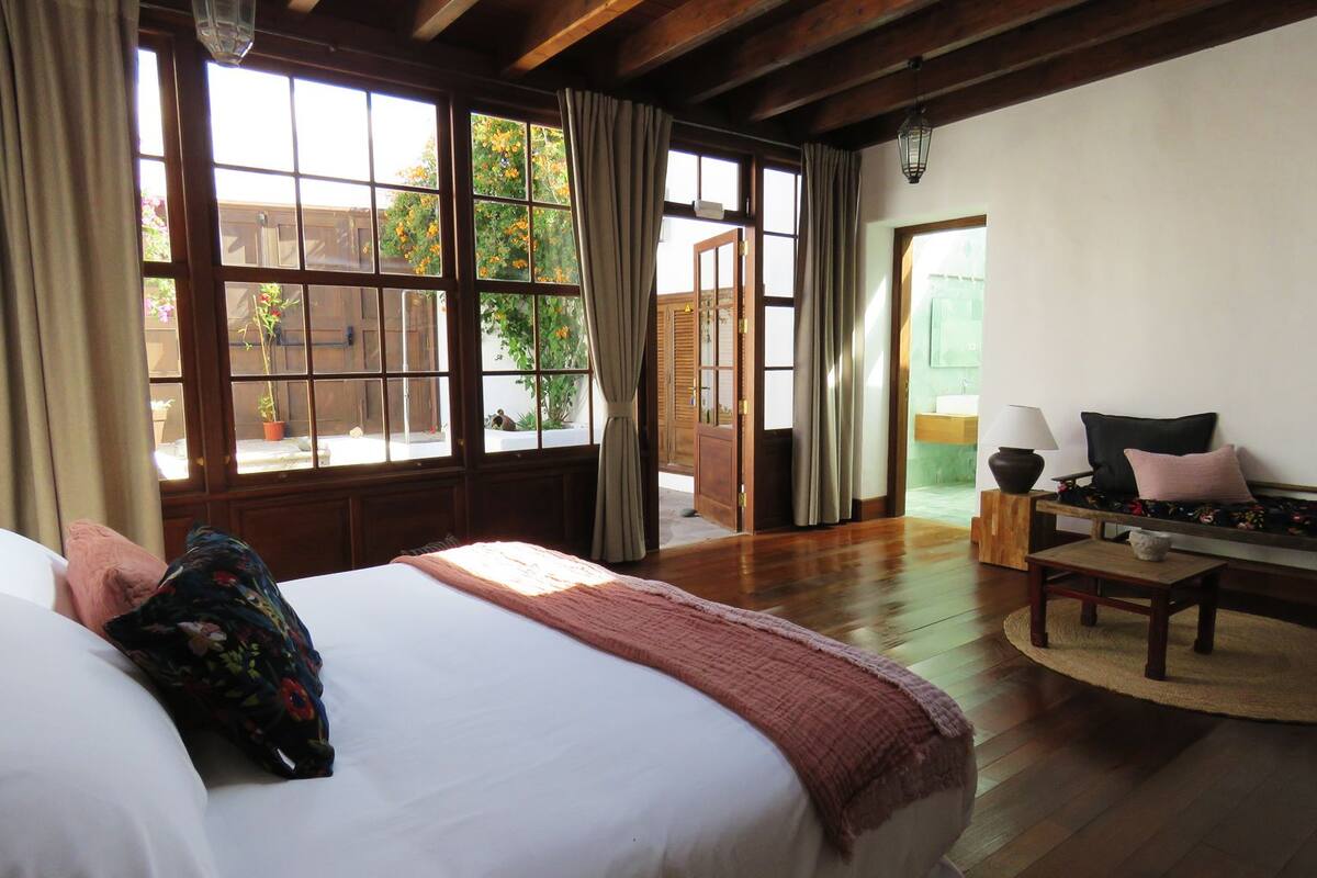 A bedroom with a large bed, a wooden bench, a small table, and a view of the outside through large windows.