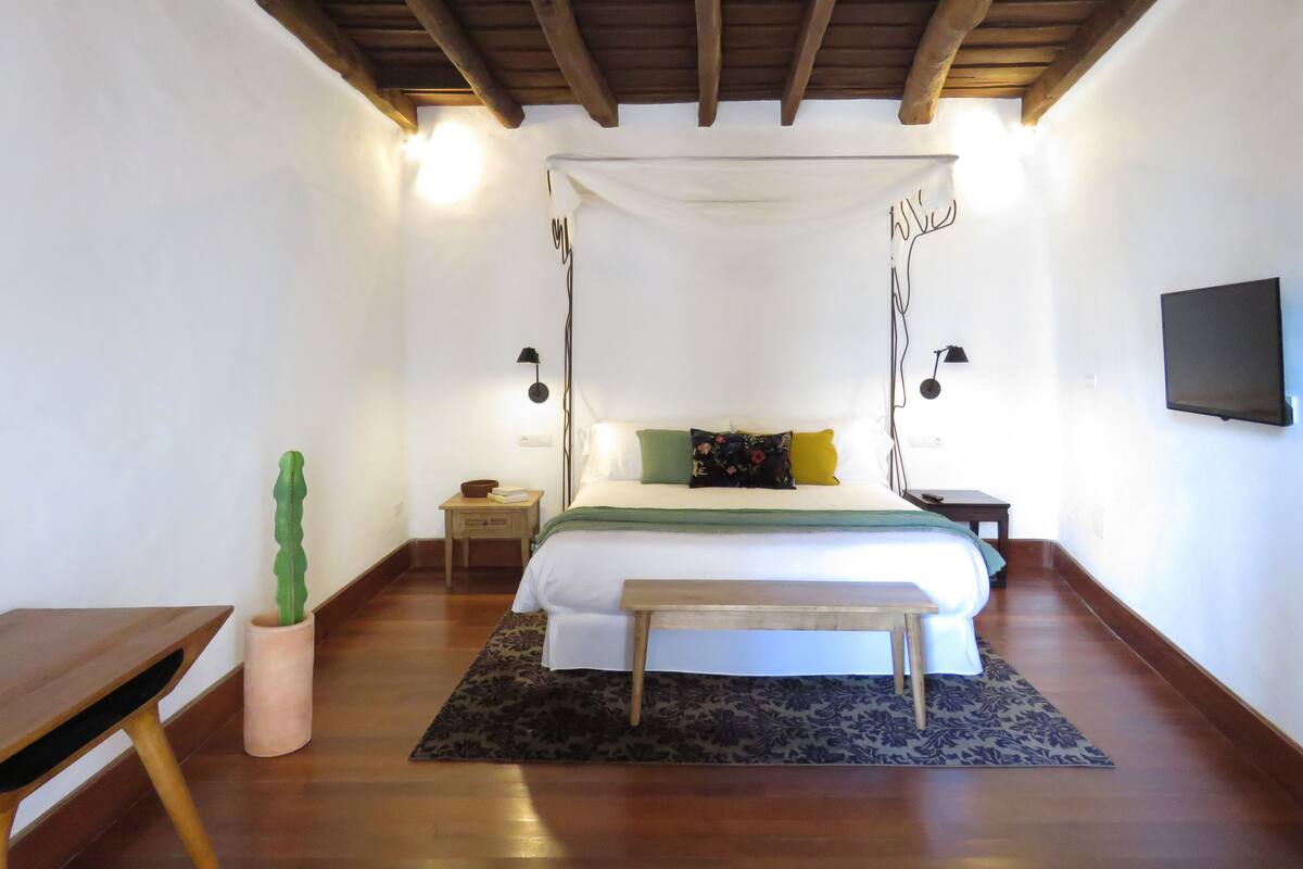 A bedroom with a canopy bed, wooden floor, bedside tables, a small table, a potted plant, and a wall-mounted TV.