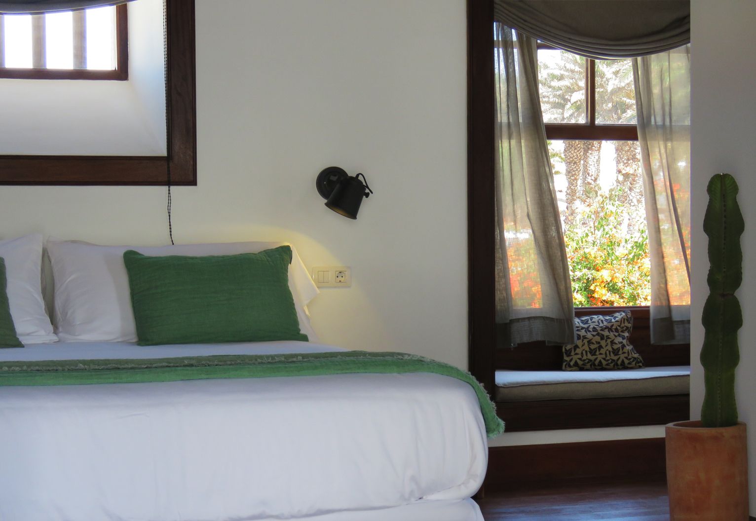 Un dormitorio con cama, mesita de noche, lámpara, ventana con cortinas y una planta de cactus.