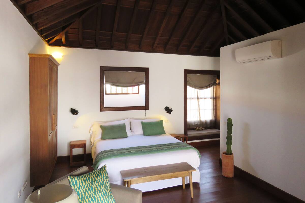 A bedroom with a wooden ceiling, a bed with green and white bedding, a wooden bench, a small table, and a potted plant.