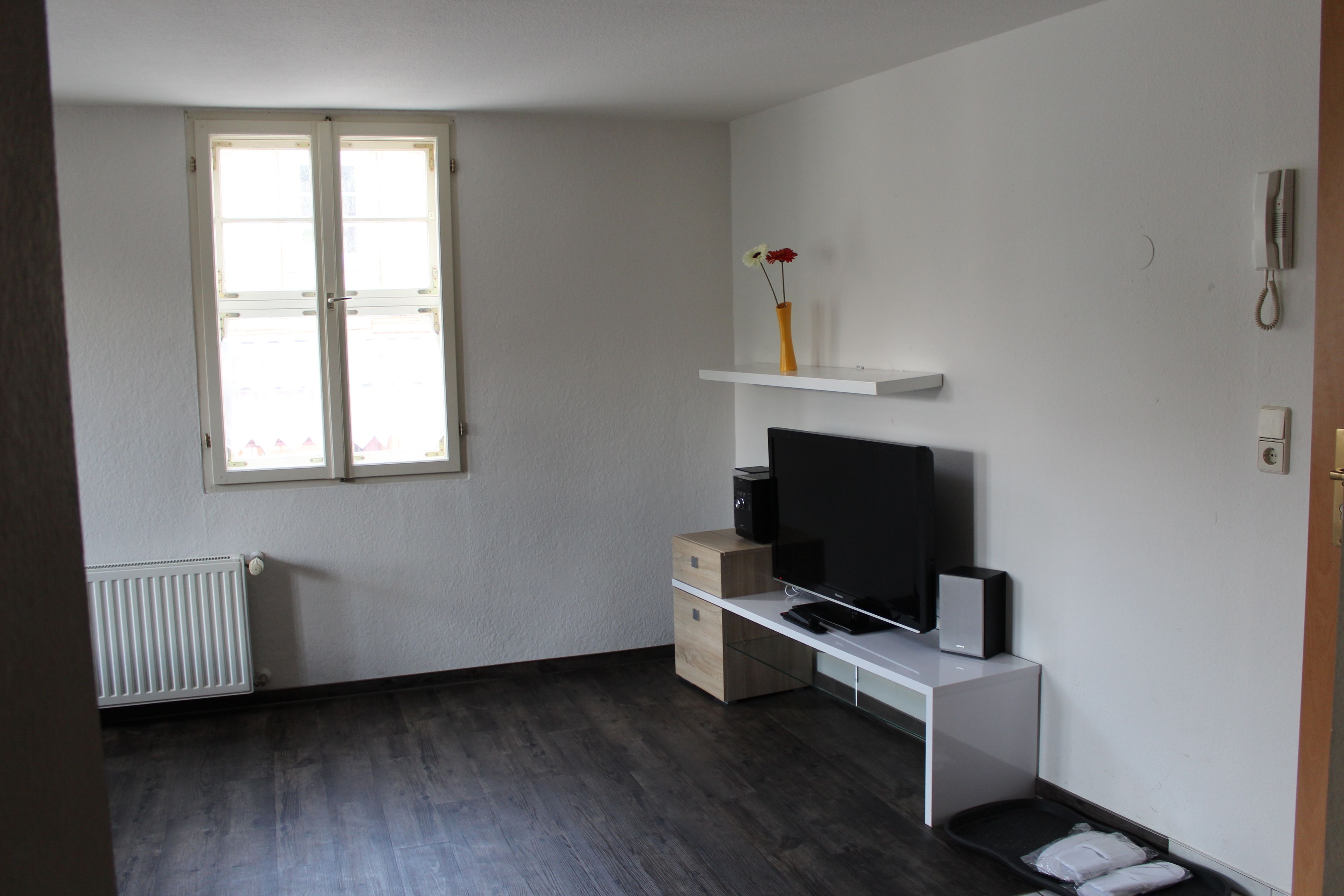 A room with a white desk, a television, a radiator, and a window with blinds.