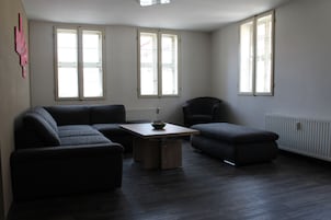 Ein modernes Wohnzimmer mit Ecksofa, einem kleinen Tisch und einem Sessel. Große Fenster sorgen für natürliches Licht.
