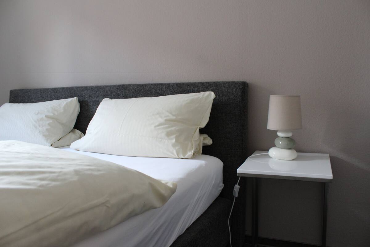 A neatly made bed with white linens and pillows, a bedside table with a lamp, and a power outlet.