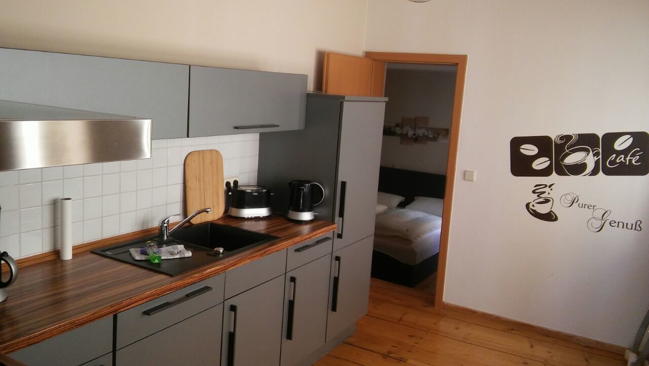 Een compacte keuken met grijze kastjes, een houten aanrecht, een spoelbak, een koffiezetapparaat en een koelkast. Een bed is zichtbaar in de aangrenzende kamer.