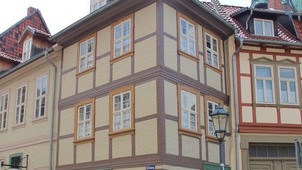 Un bâtiment traditionnel à colombages, avec une porte et des fenêtres vertes, une rue pavée et, au loin, le clocher d’une église.