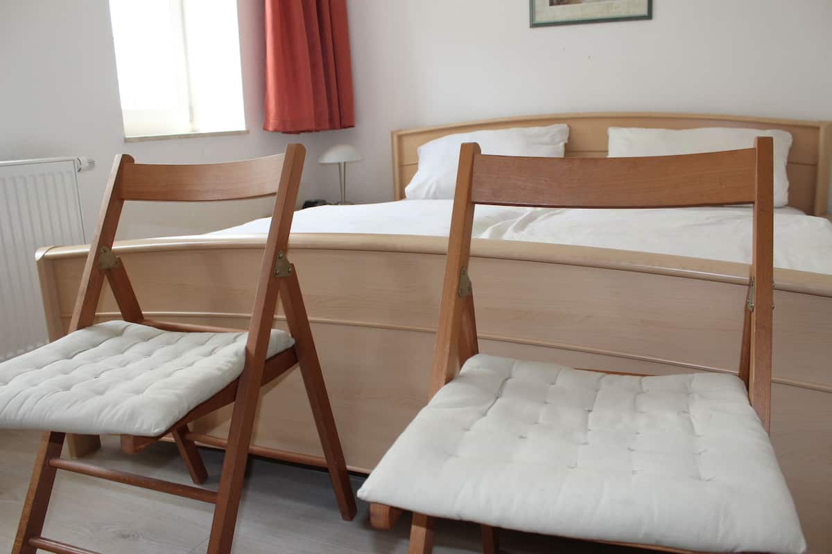 Een slaapkamer met een bed, twee houten stoelen met zachte zittingen, een radiator, een raam met gordijnen en een wandlamp.