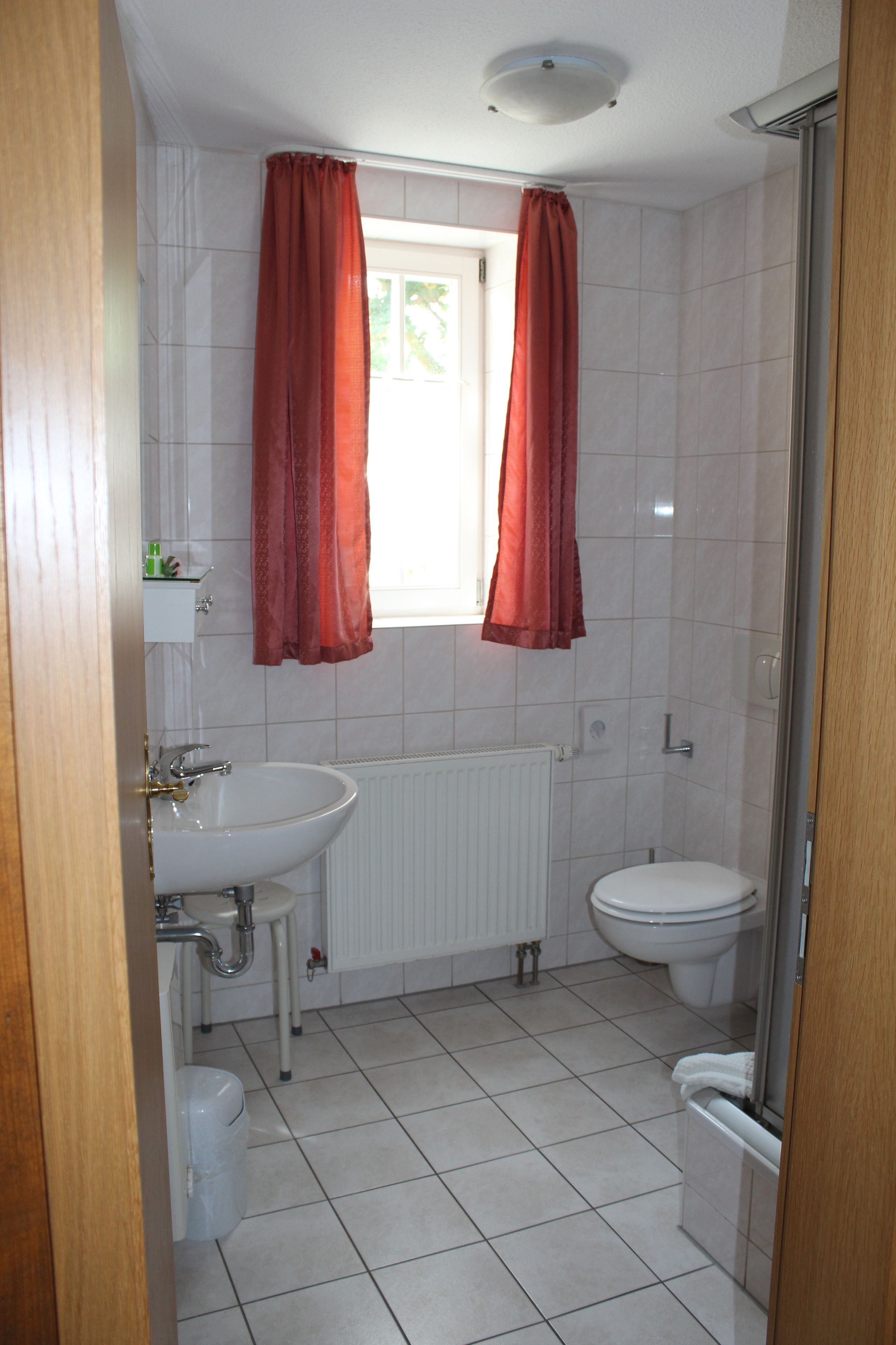 A bathroom with a white sink, toilet, and shower. There are red curtains over the window and a white radiator on the wall.