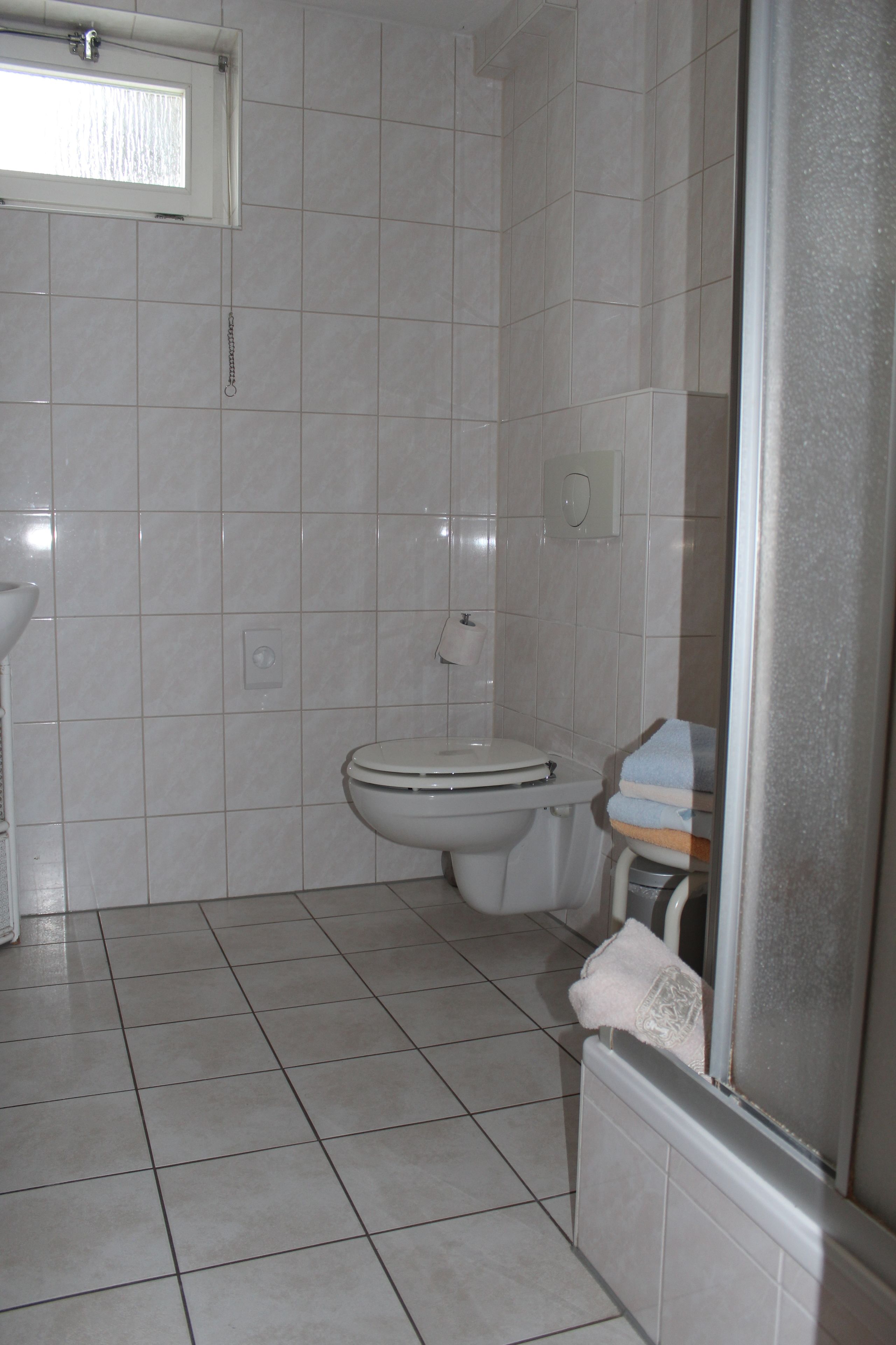 Un baño con paredes y suelo de azulejos blancos, un inodoro, un lavabo y un ventanal.