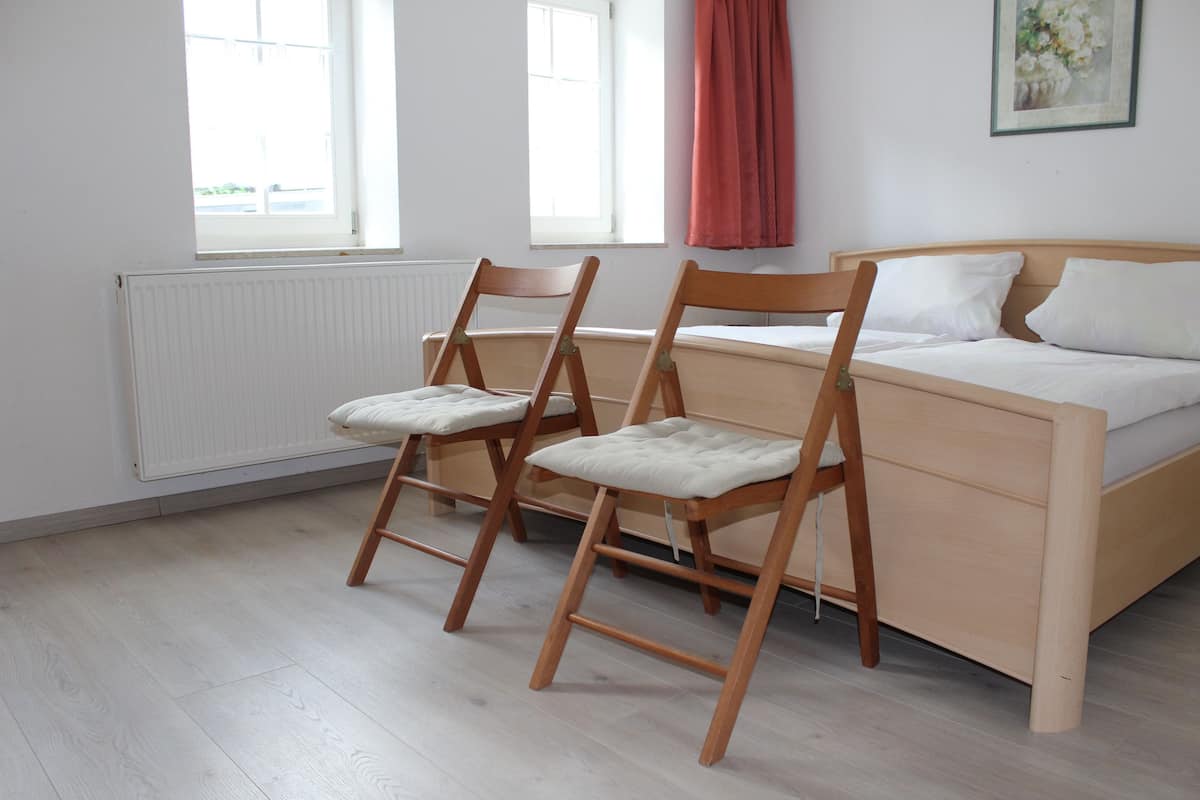 Een slaapkamer met een bed, twee houten stoelen, een radiator en ramen.