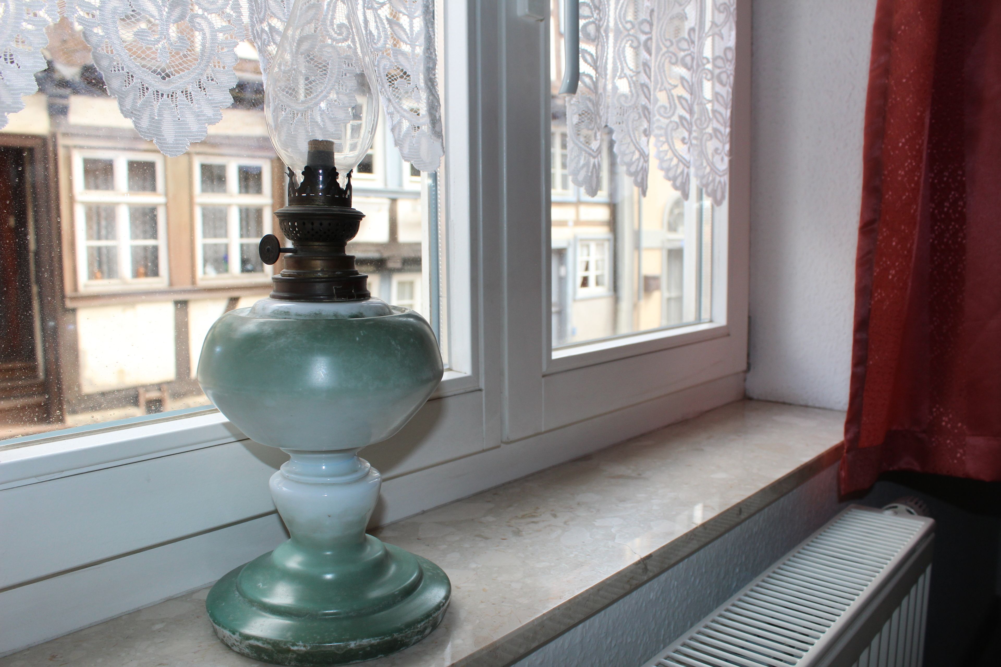 Eine alte, grüne Tischlampe auf einem Fensterbrett mit Spitzenvorhängen und Blick auf Gebäude.