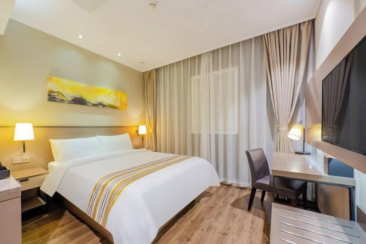 A hotel room with a bed, bedside lamps, a desk, and a large window with curtains.
