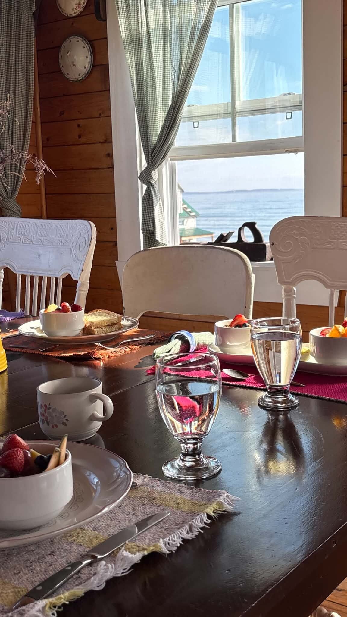 A dining table set with plates, bowls, glasses, and cutlery, with a view of the sea through a window.