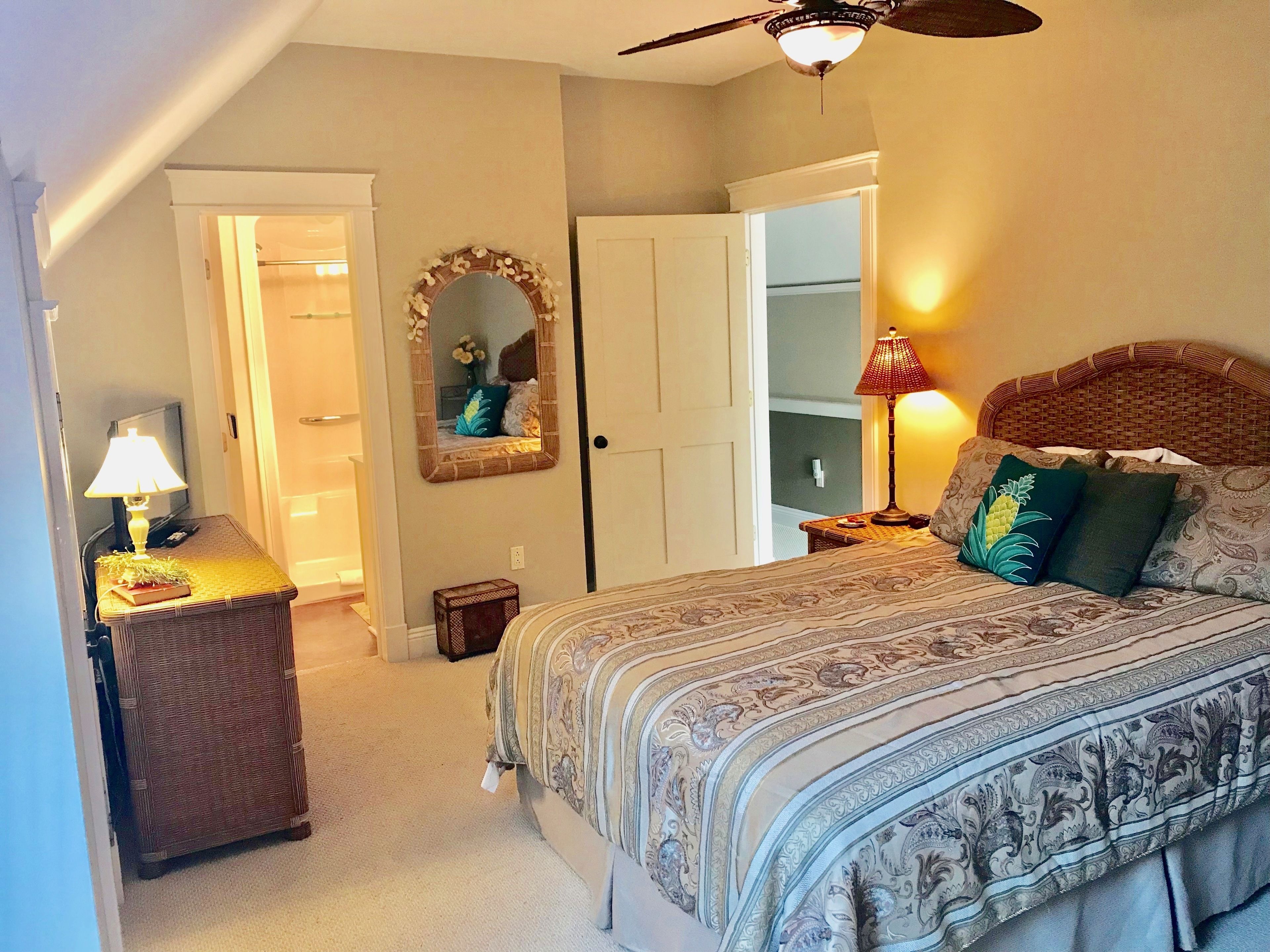 A bedroom with a large bed, a nightstand with a lamp, a mirror, and a ceiling fan.