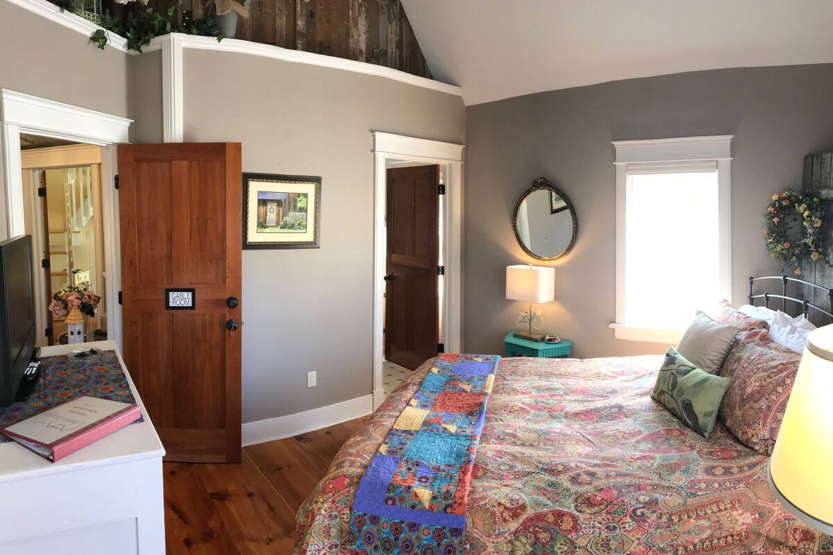 Een slaapkamer met een groot bed, een nachtkastje, een lamp, een spiegel en een deur met de tekst 'GABLE ROOM'.