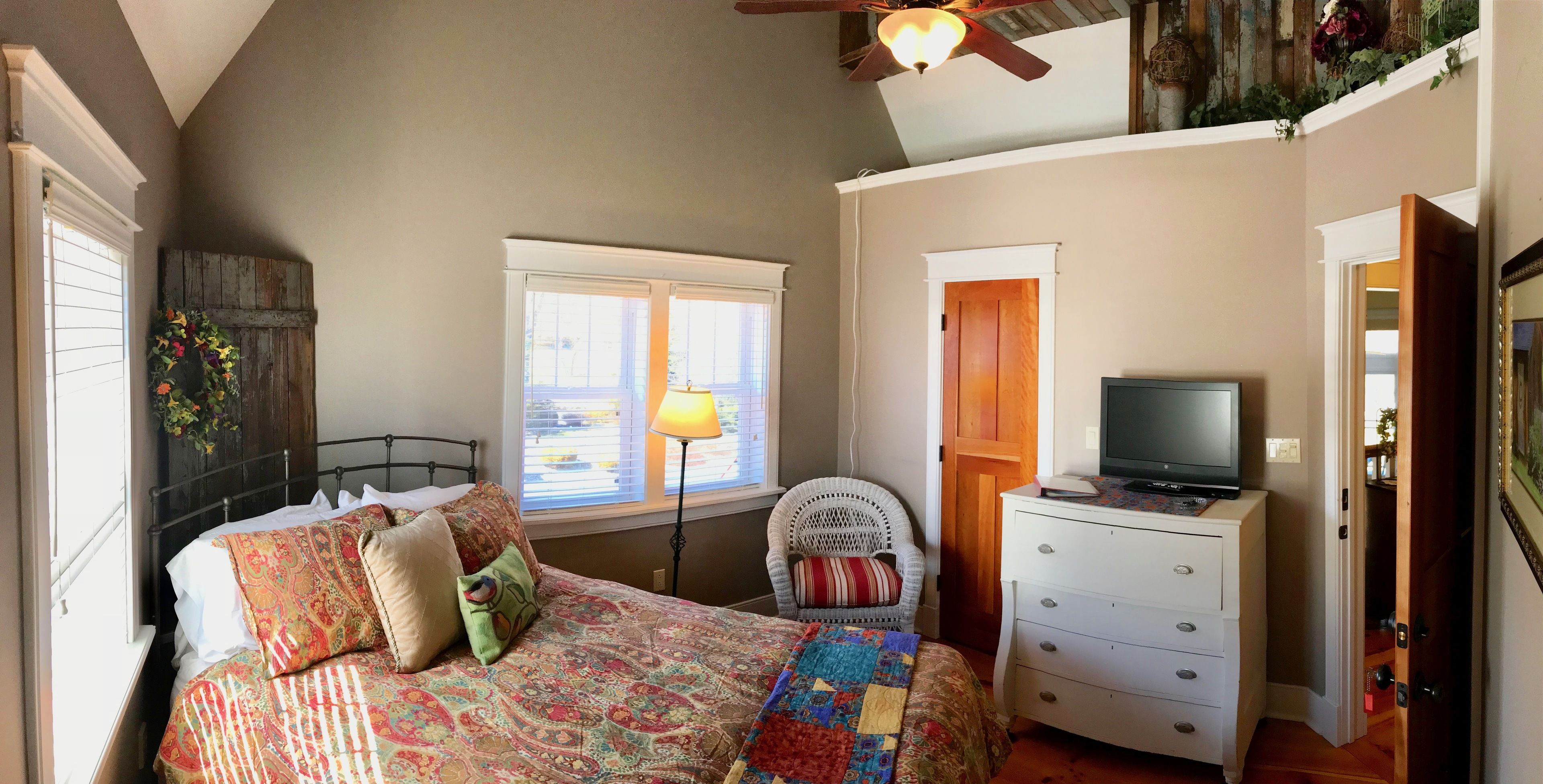 A bedroom with a bed, a dresser, a television, a window with blinds, and a ceiling fan.
