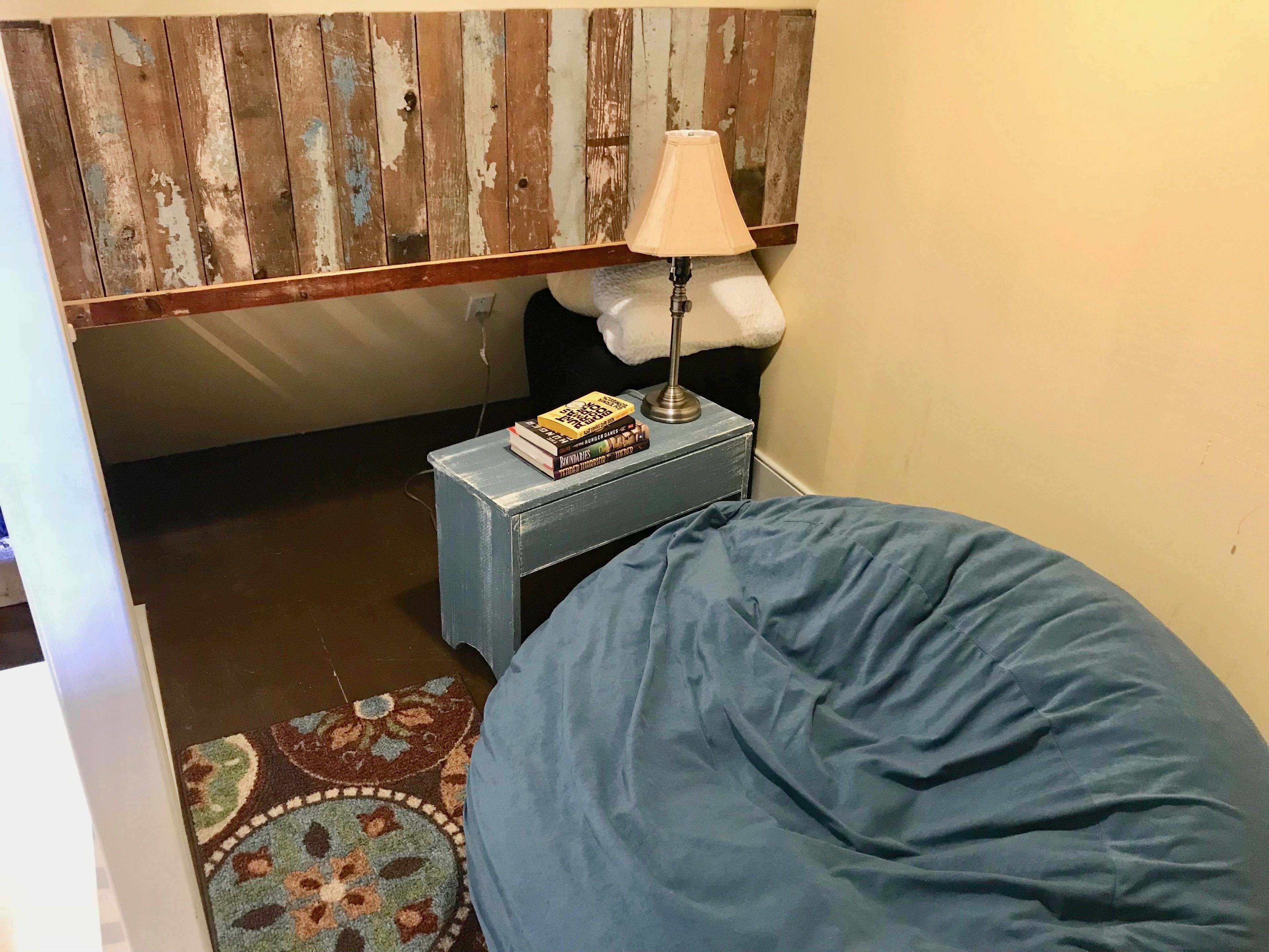 A bedroom with a blue bed, a small bedside table with a lamp and books, a wooden headboard, and a patterned rug.