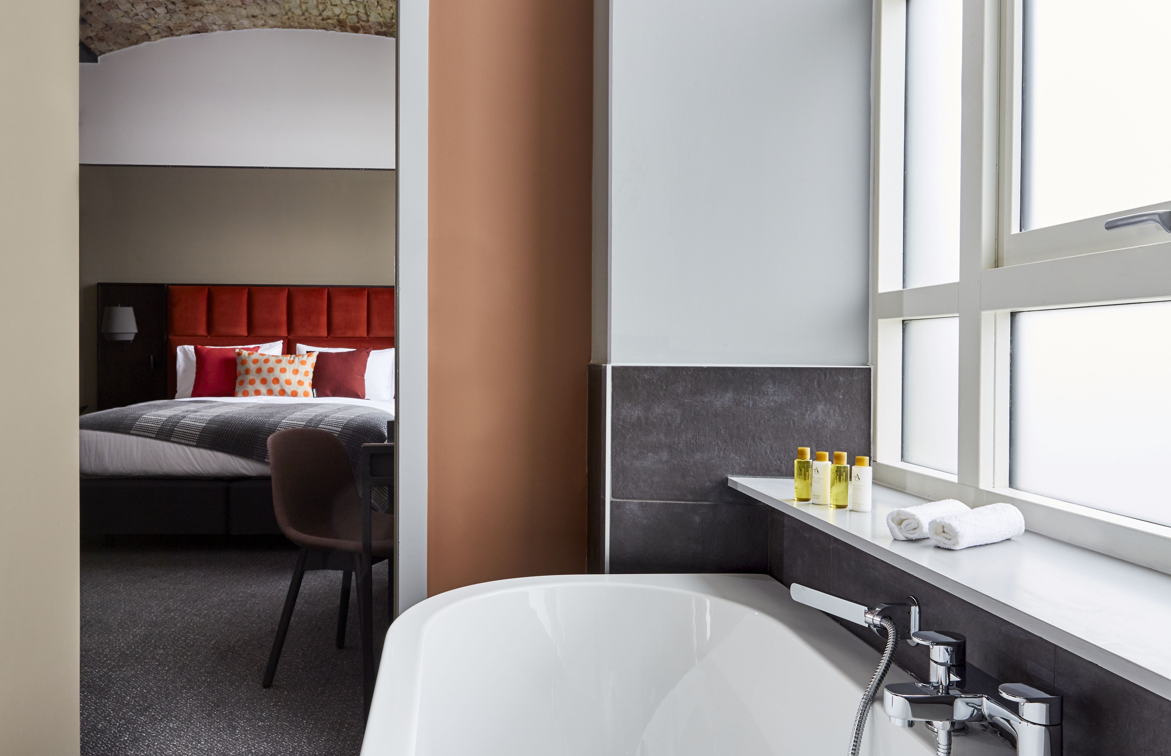 Moderni hotellihuone, jossa on sänky, tuoli, kylpyamme ja hygieniatarvikkeet ikkunalaudalla.