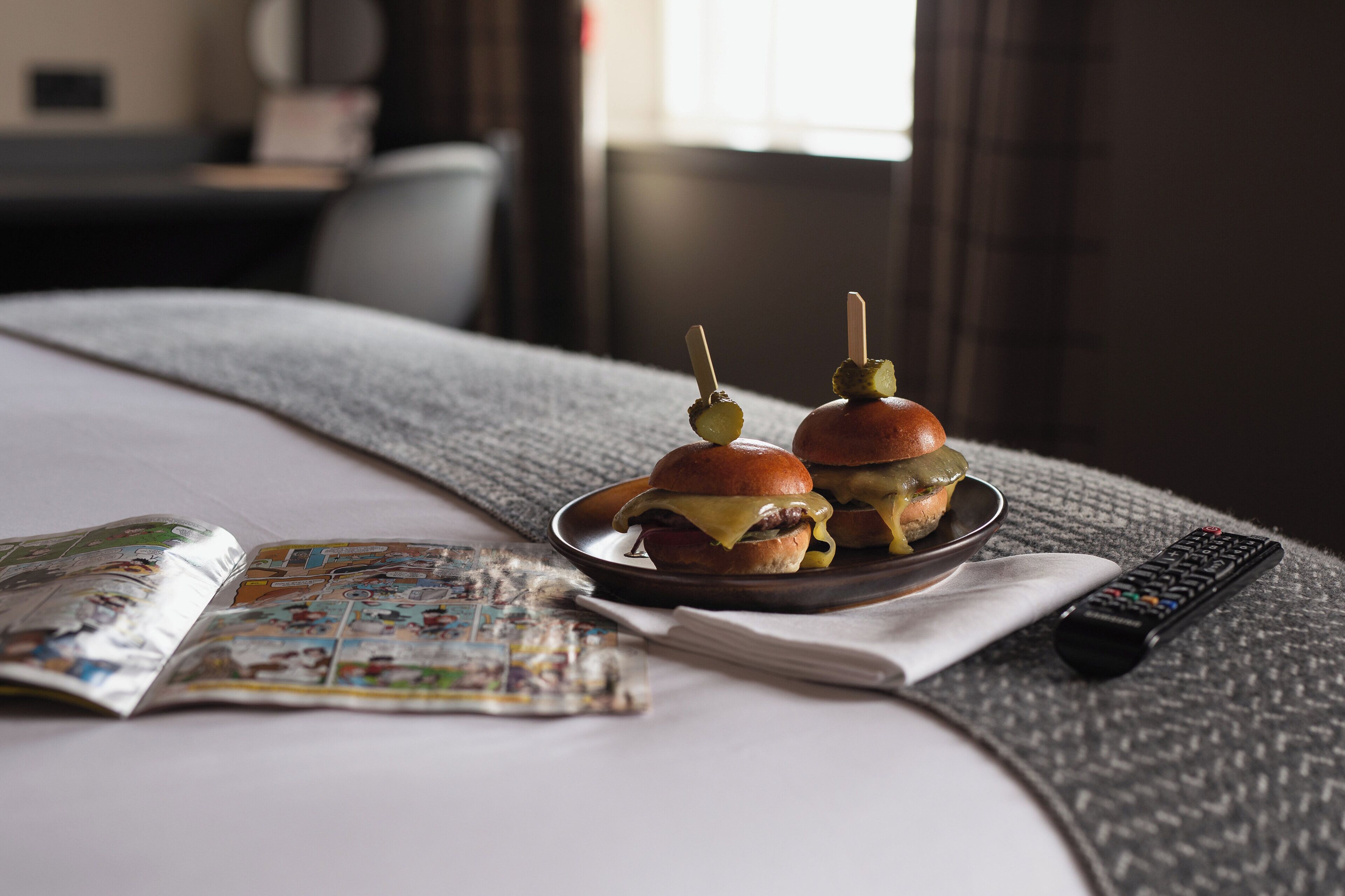A bed with a blanket, a plate with two burgers, a remote control, and a magazine.