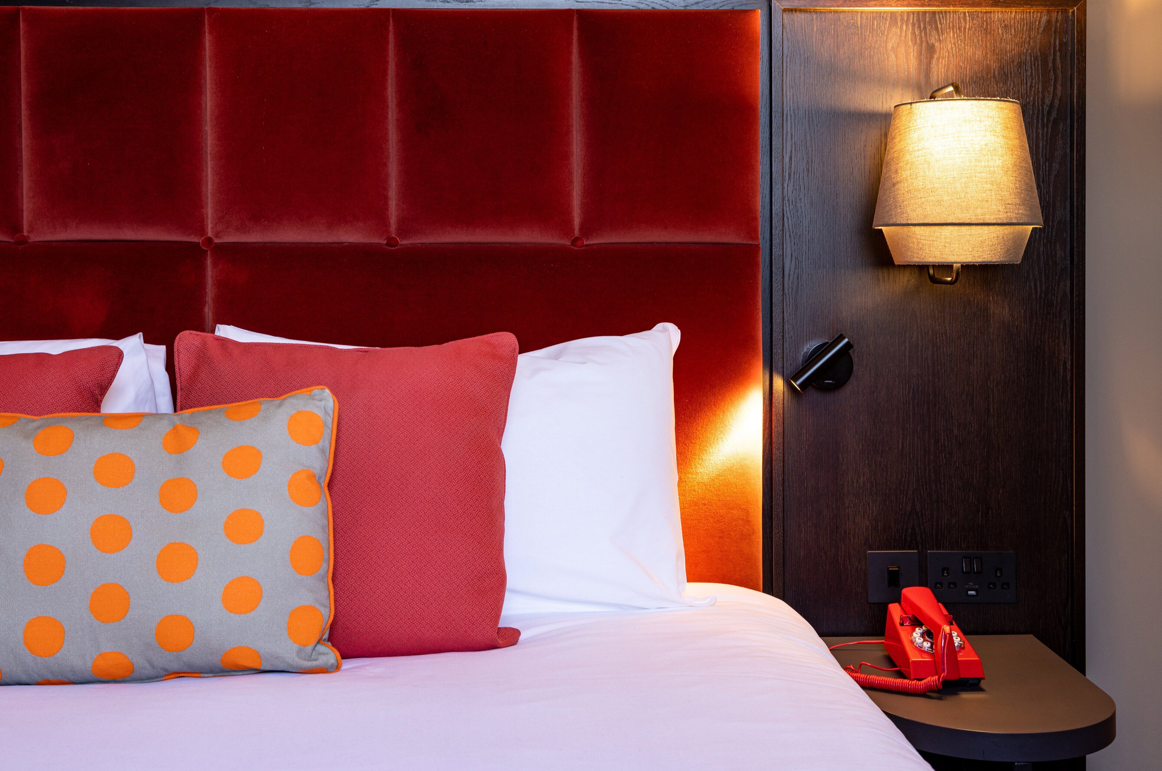 A hotel room with a red upholstered headboard, a bed with white and red pillows, a gray pillow with orange polka dots, and a red telephone on a bedside table.