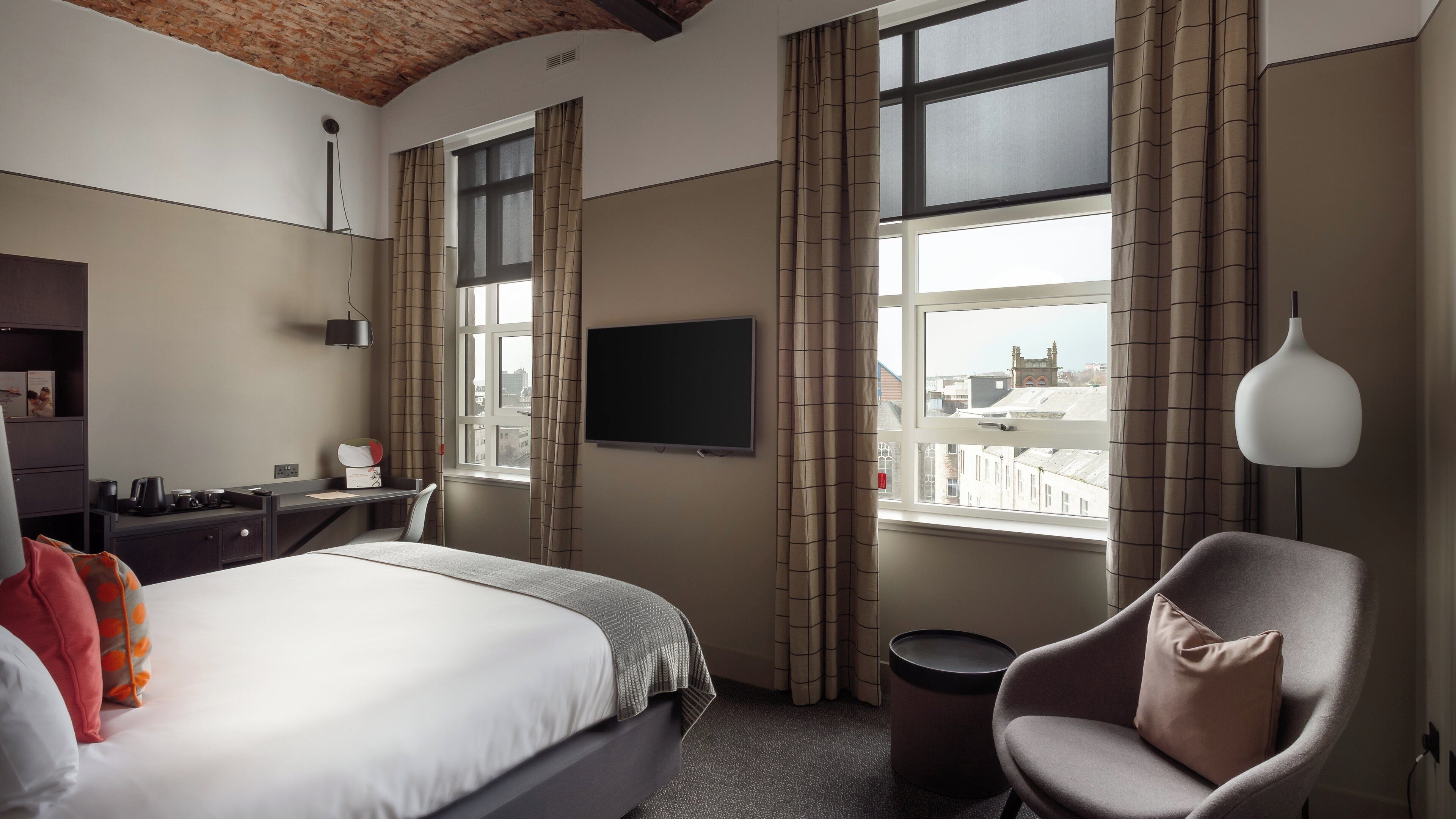 A hotel room with a bed, desk, chair, TV, and large windows with curtains.