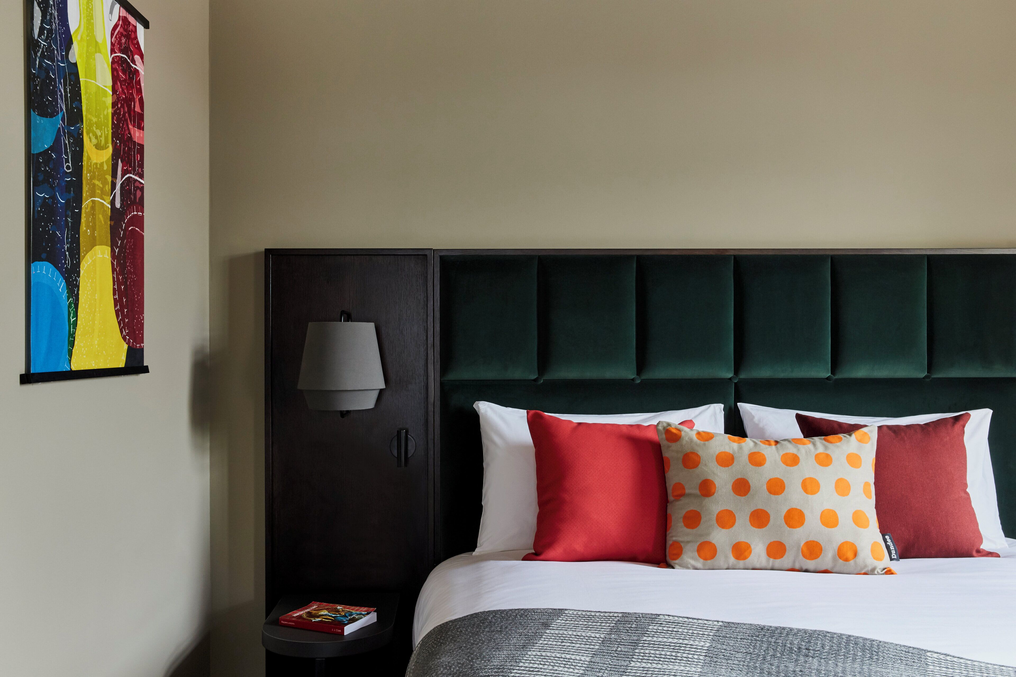 A hotel room with a bed, a nightstand, a lamp, and a colorful artwork on the wall.