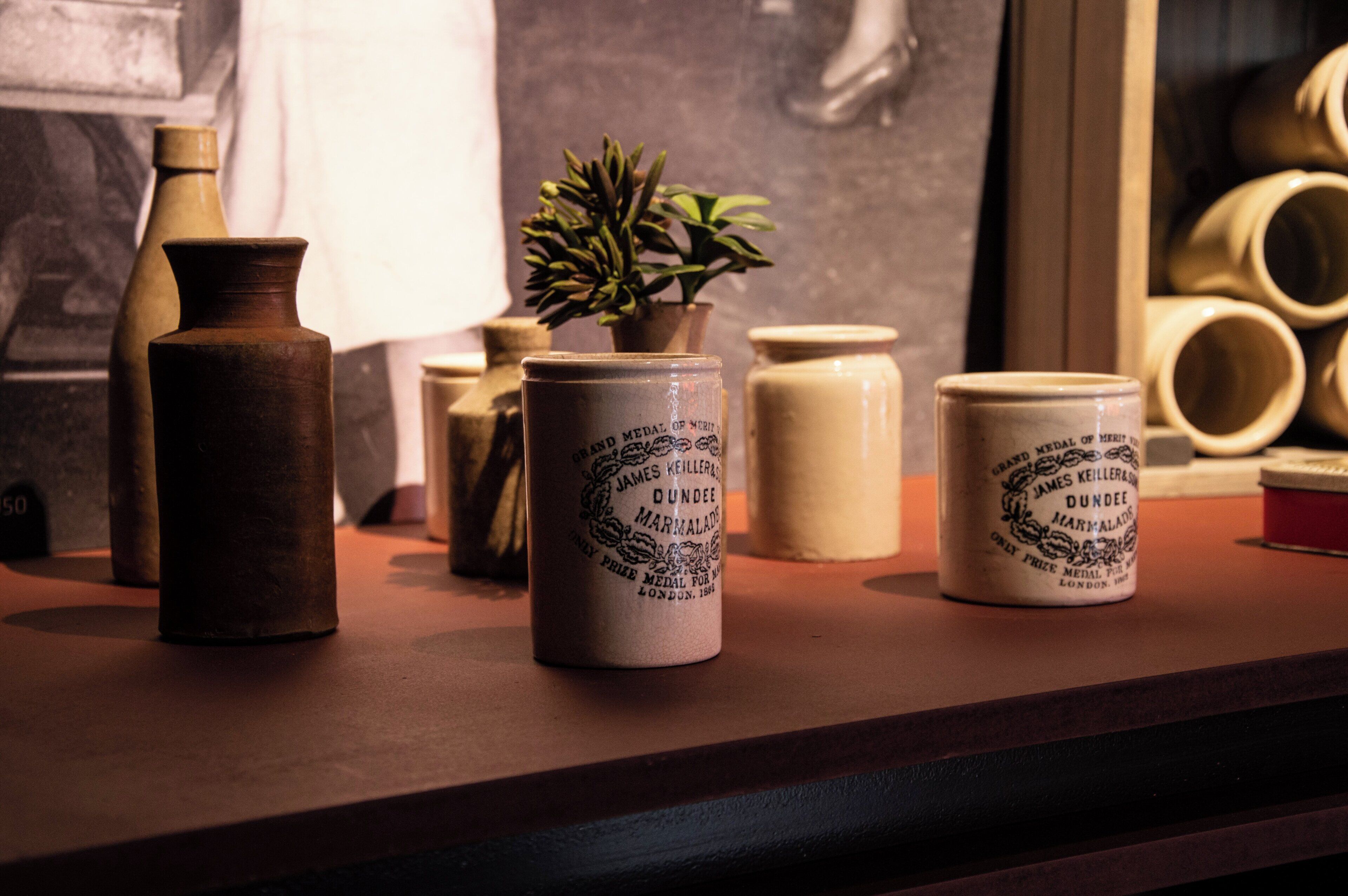 A display of ceramic items including vases and jars with inscriptions, a potted plant, and a wooden object on a shelf.