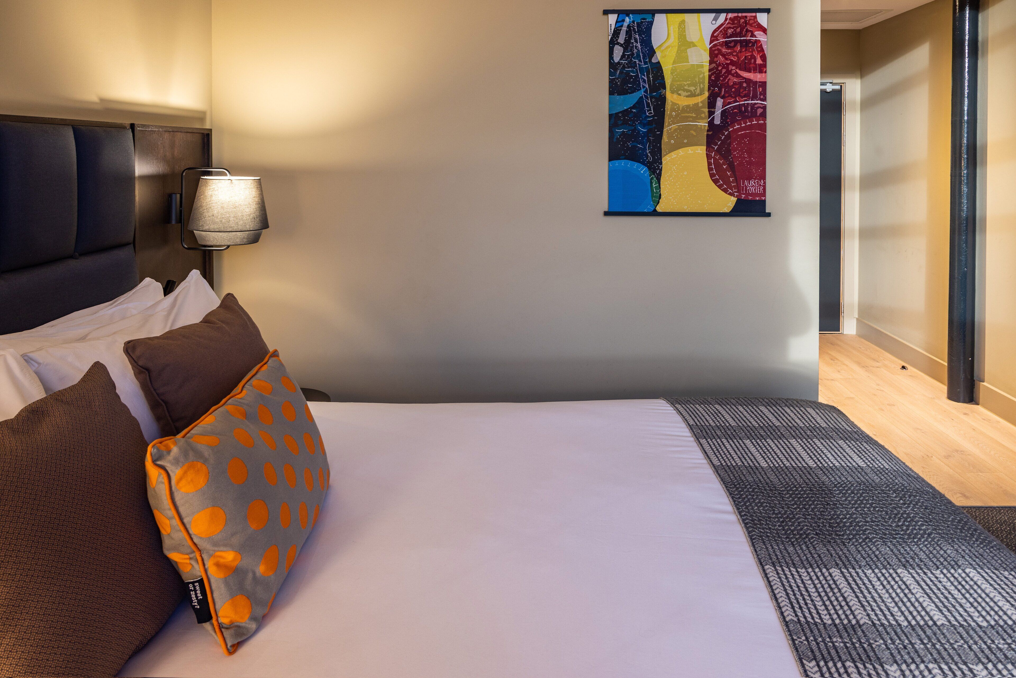 Una habitación de hotel con una cama bien hecha, una lámpara de pared y un cuadro abstracto y colorido.