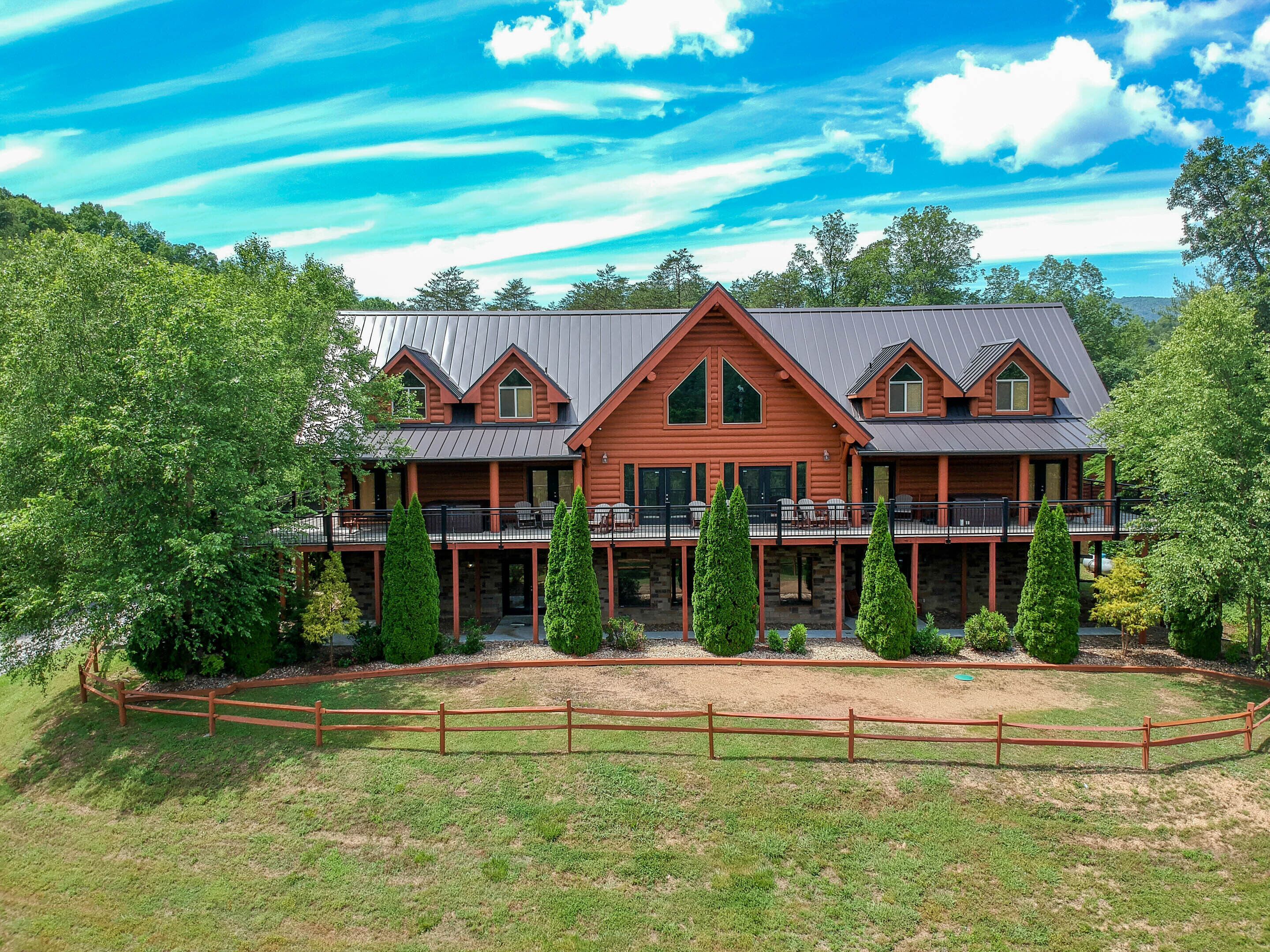 A large, two-story log cabin with a balcony and multiple windows, surrounded by lush greenery and a fenced yard.