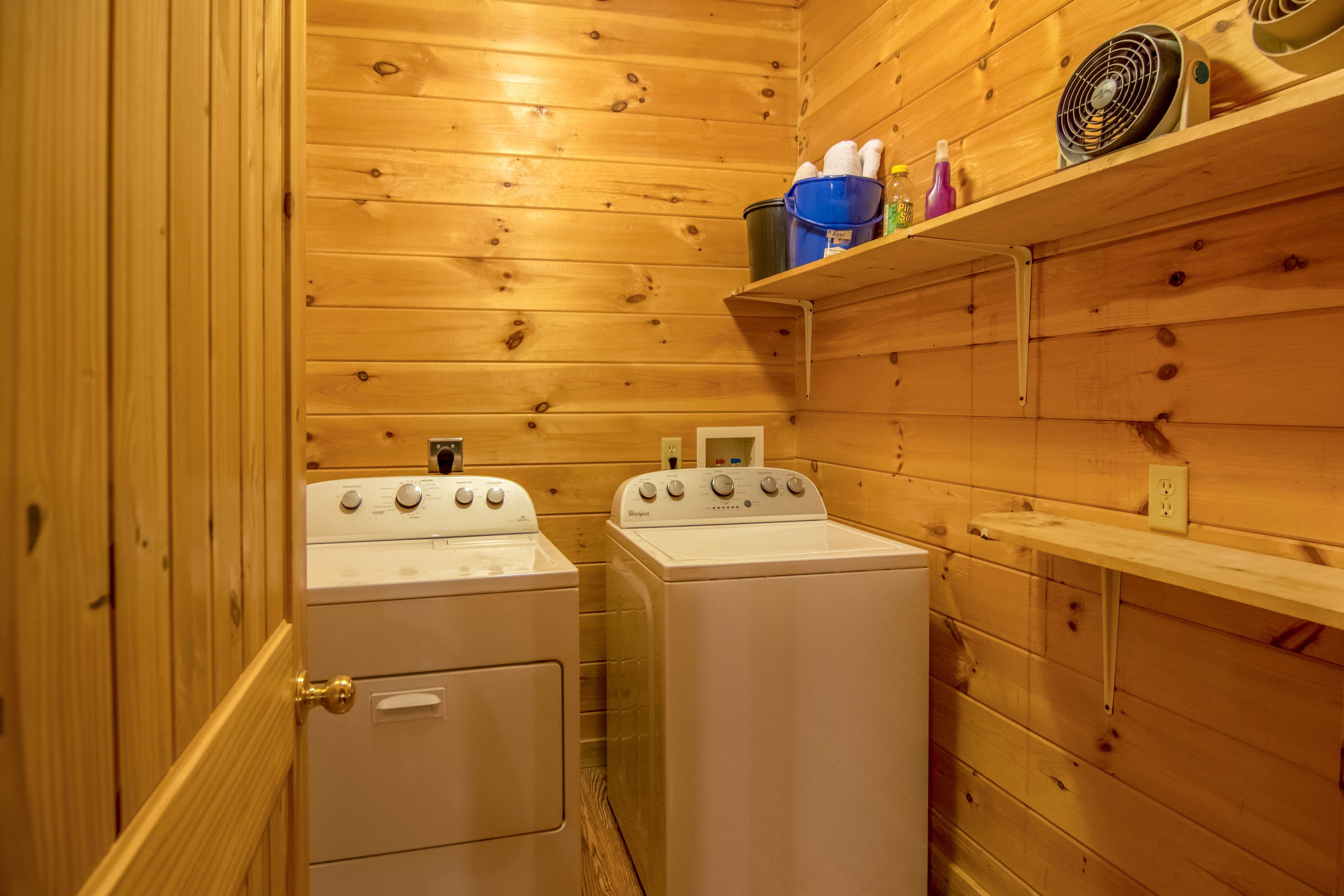 A compact laundry room with two white washers and dryers, wooden walls, and a shelf with cleaning supplies.