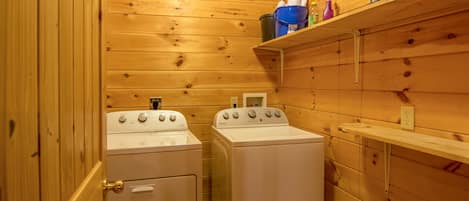A compact laundry room with two white washers and dryers, wooden walls, and a shelf with cleaning supplies.