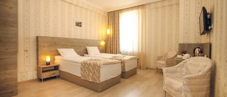 A hotel room with two beds, a wooden headboard, a nightstand with a lamp, a chair, a small table with a mirror, and a window with curtains.