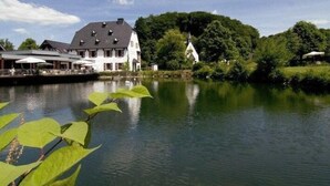 A lakeside hotel with a large building, outdoor seating area, and a small church nearby. - Malteser Komturei Hotel / Restaurant (Bergisch Gladbach)